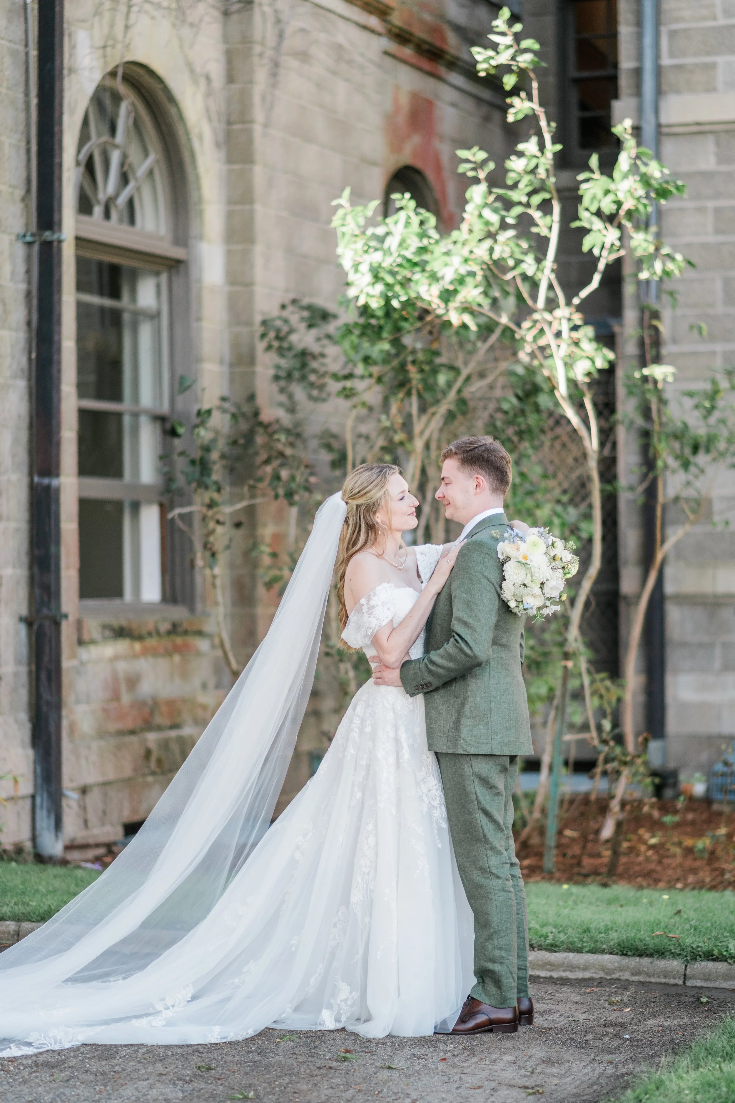 A bride and groom sharing a moment outside, with the bride in a white gown and veil, and the groom in a green suit holding a bouquet of flowers, standing near an old stone building with trees in the background.