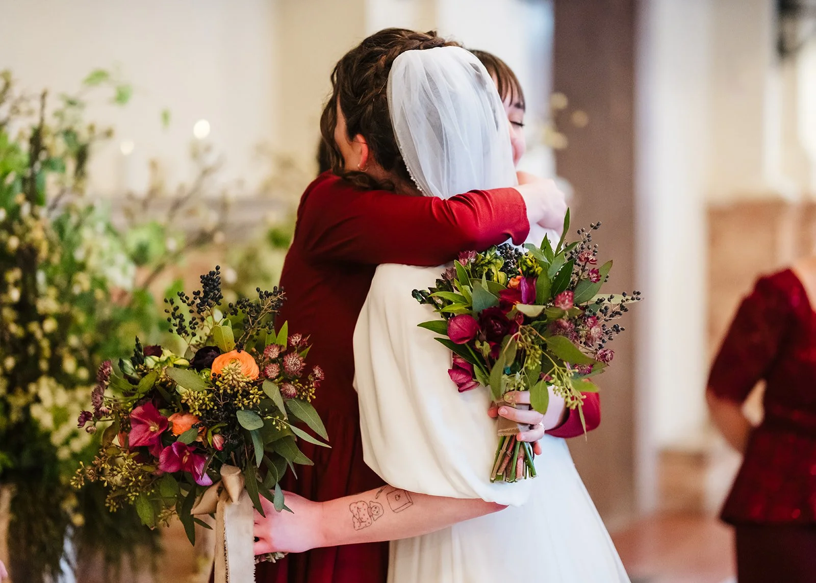 Two women hugging at a wedding, one in a white dress, holding a bouquet, the other in a burgundy dress, with a tattoo of a dog on her forearm.