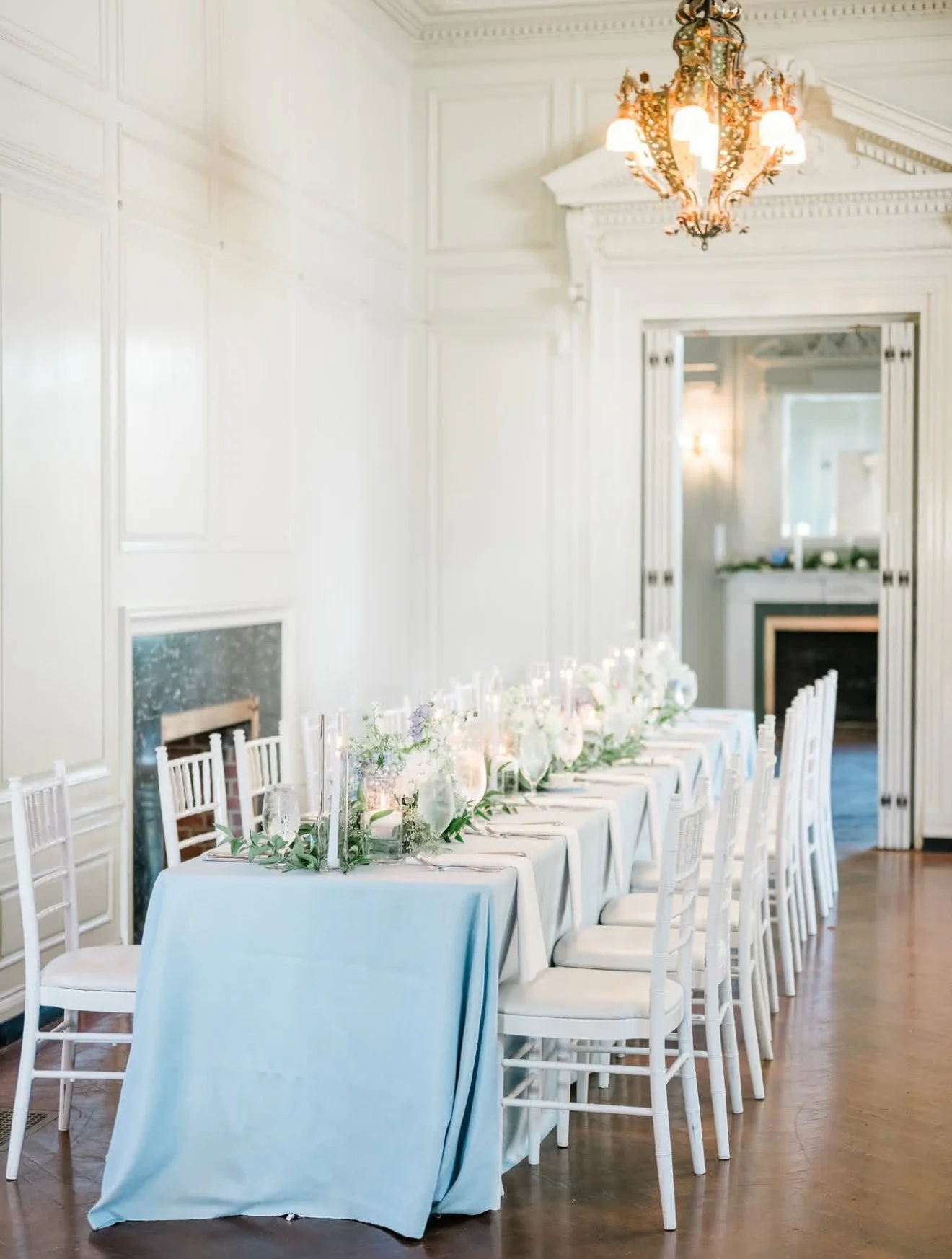 Elegant dining room with a long table decorated with floral centerpieces, candles, and white chairs, lit by a large ornate chandelier.