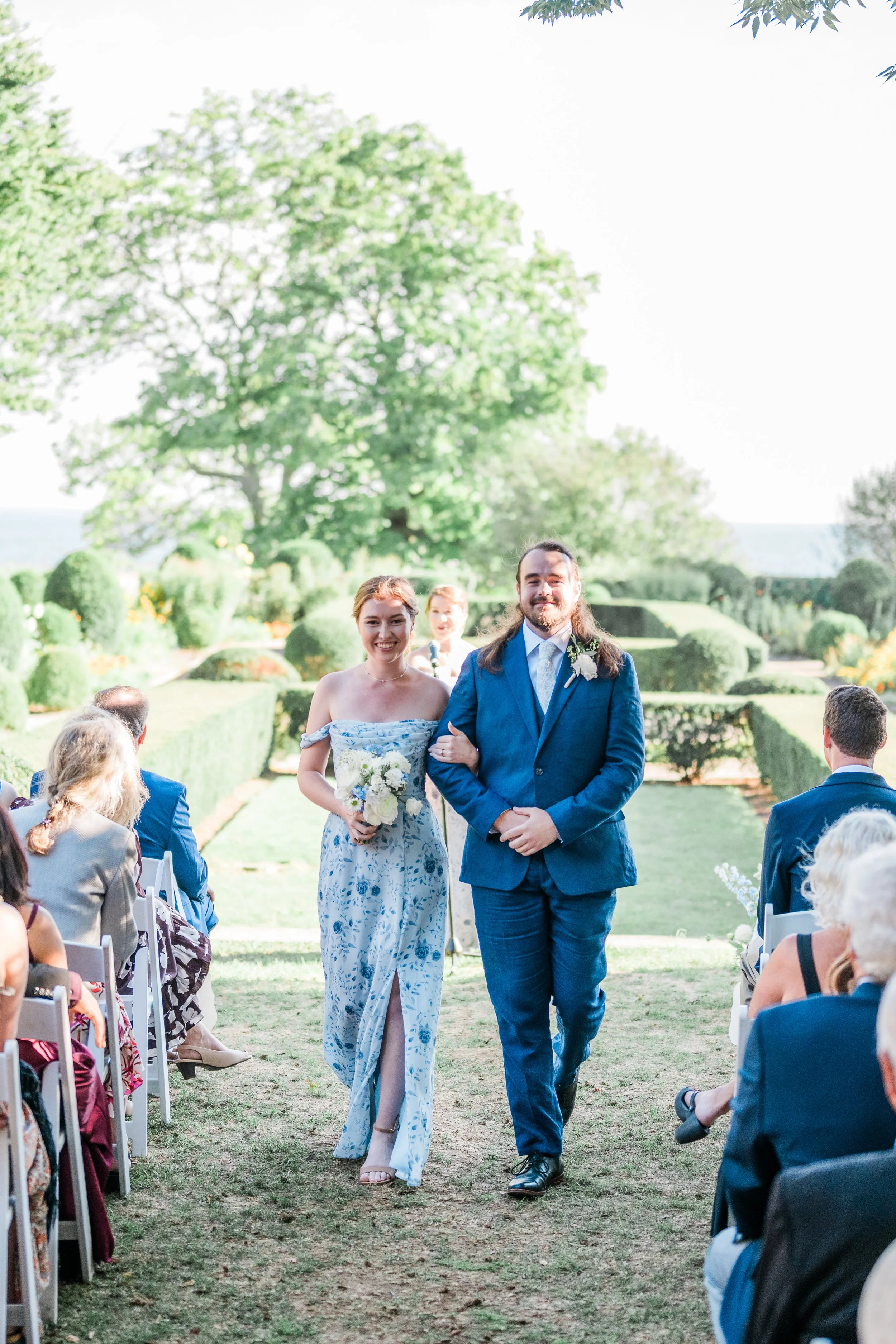 Couple walking down the aisle at a wedding ceremony outdoors, surrounded by guests seated on both sides, lush greenery, and clear sky.