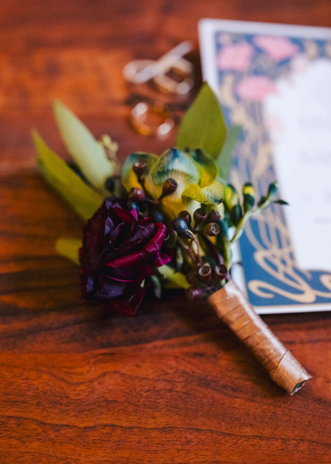 A boutonniere with green leaves, dark red flower, and small berries, resting on a wooden surface with blurred jewelry and a card in the background.