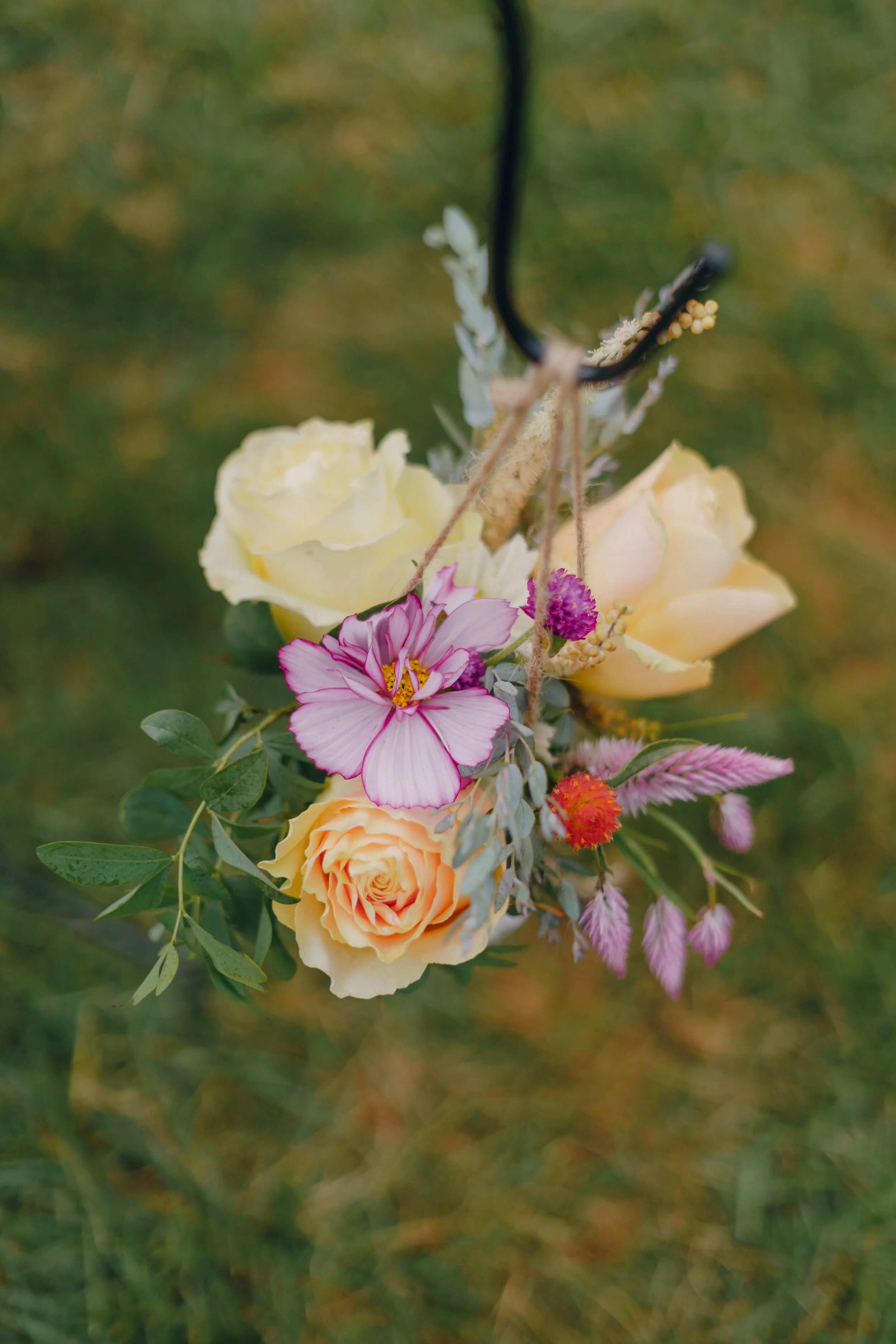 A hanging bouquet of mixed flowers including yellow roses, pink and purple blossoms, and green foliage, suspended from a black hook.