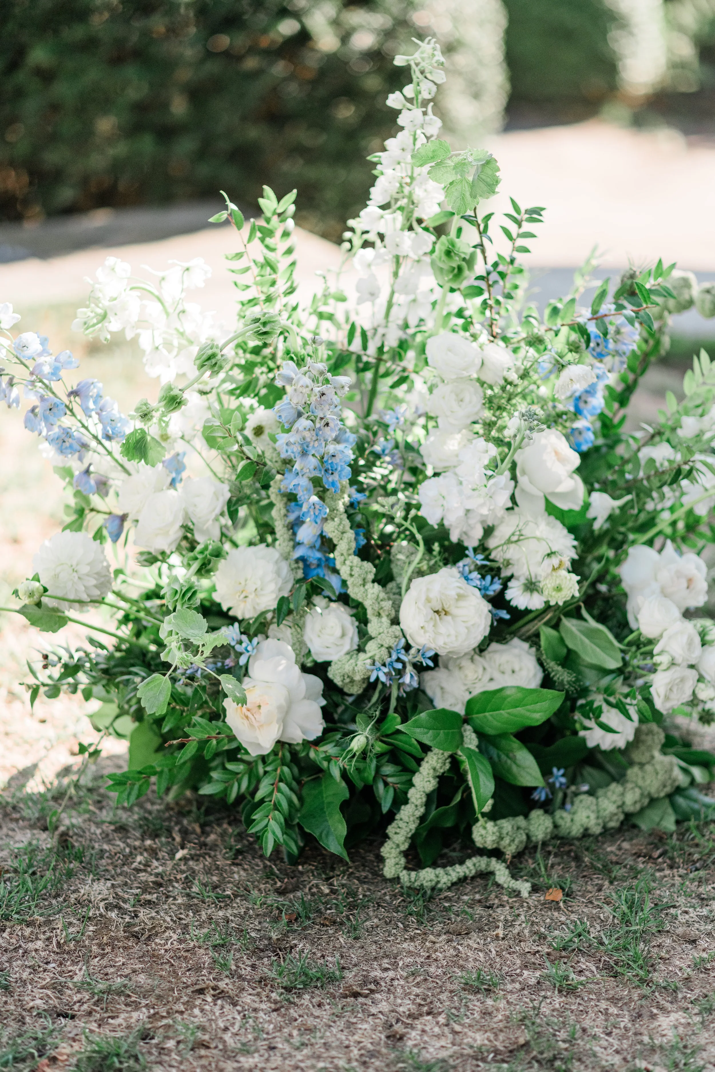 A large floral arrangement with white and light blue flowers and green foliage resting on a patch of dirt and grass outdoors.