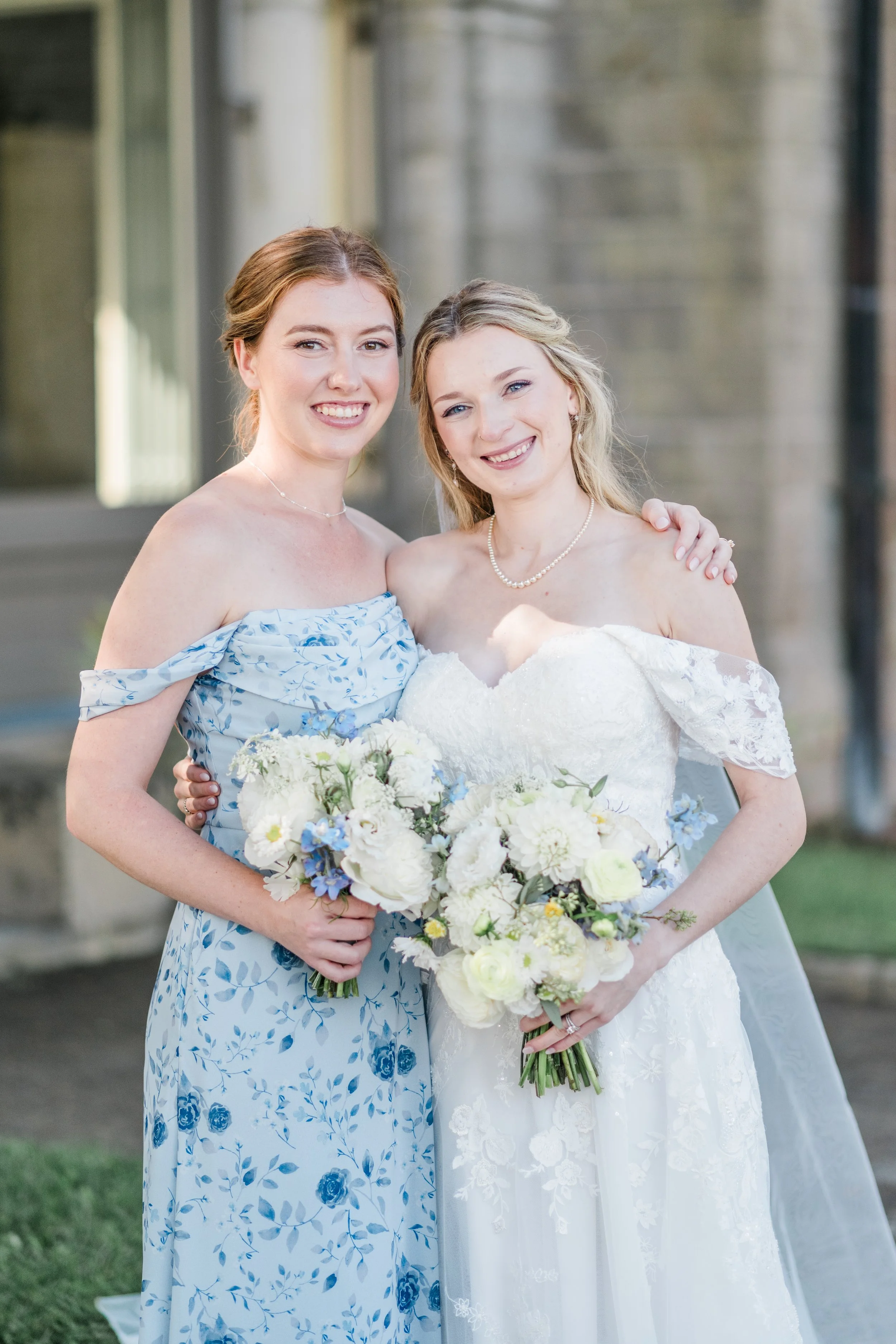 Two women smiling and posing outdoors, one in a wedding dress and the other in a floral dress, holding bouquets of white and blue flowers.