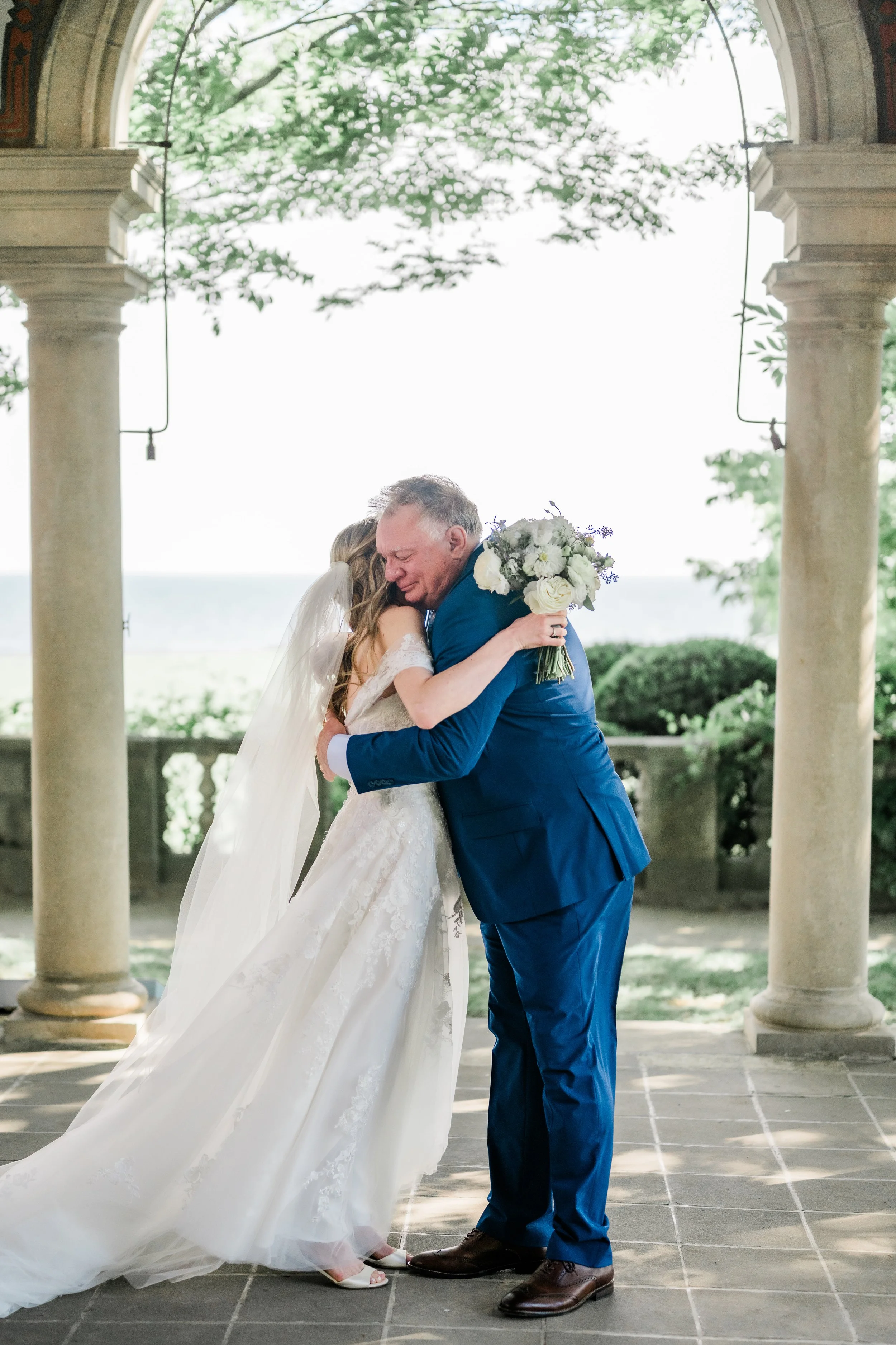 A bride in a white wedding gown and veil hugging an older man in a blue suit who is holding a bouquet of white and purple flowers, on a stone patio with columns and greenery in the background.