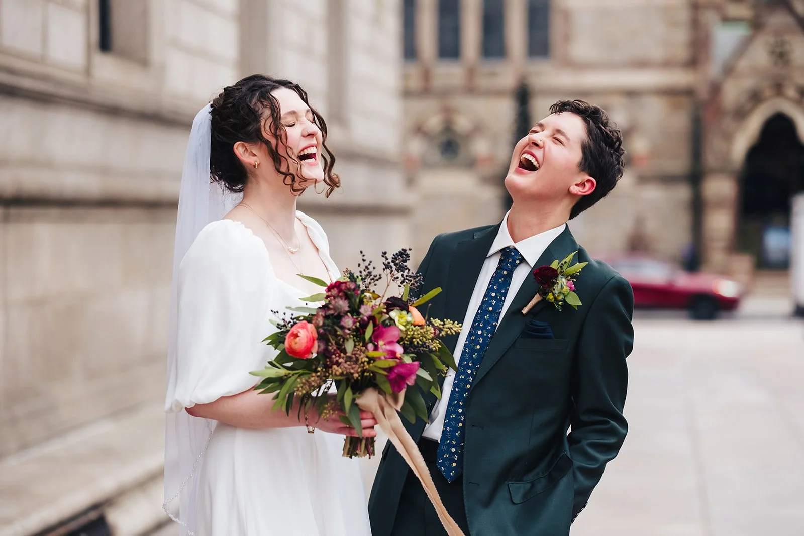 A bride and groom laughing together outdoors, with the bride holding a bouquet of colorful flowers and the groom in a dark suit with a boutonniere.