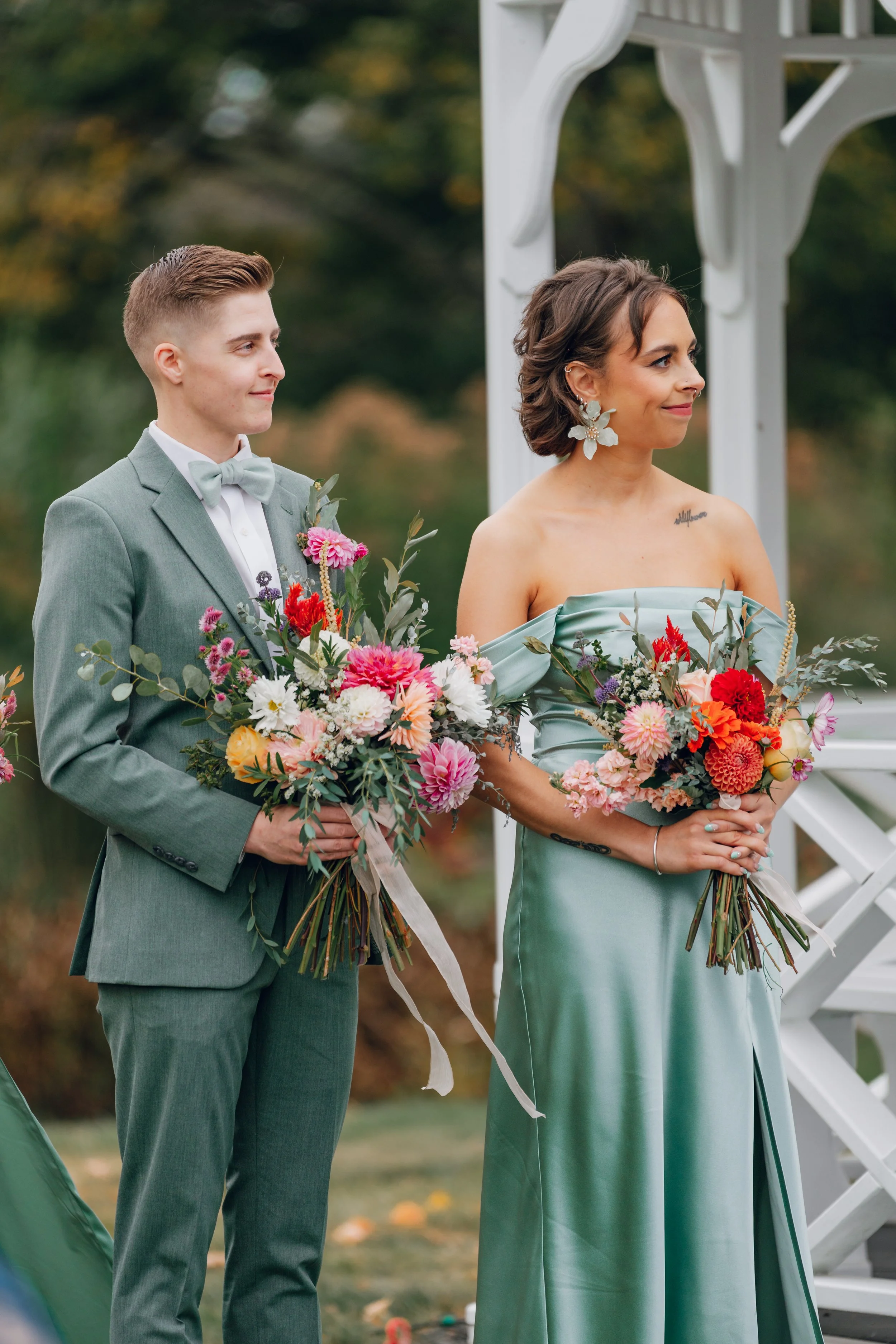 A couple at their wedding ceremony standing outdoors in front of a white arch, holding colorful flower bouquets. The groom is in a light gray suit with a gray bow tie, and the bride is in a pastel blue off-the-shoulder dress with large floral earring