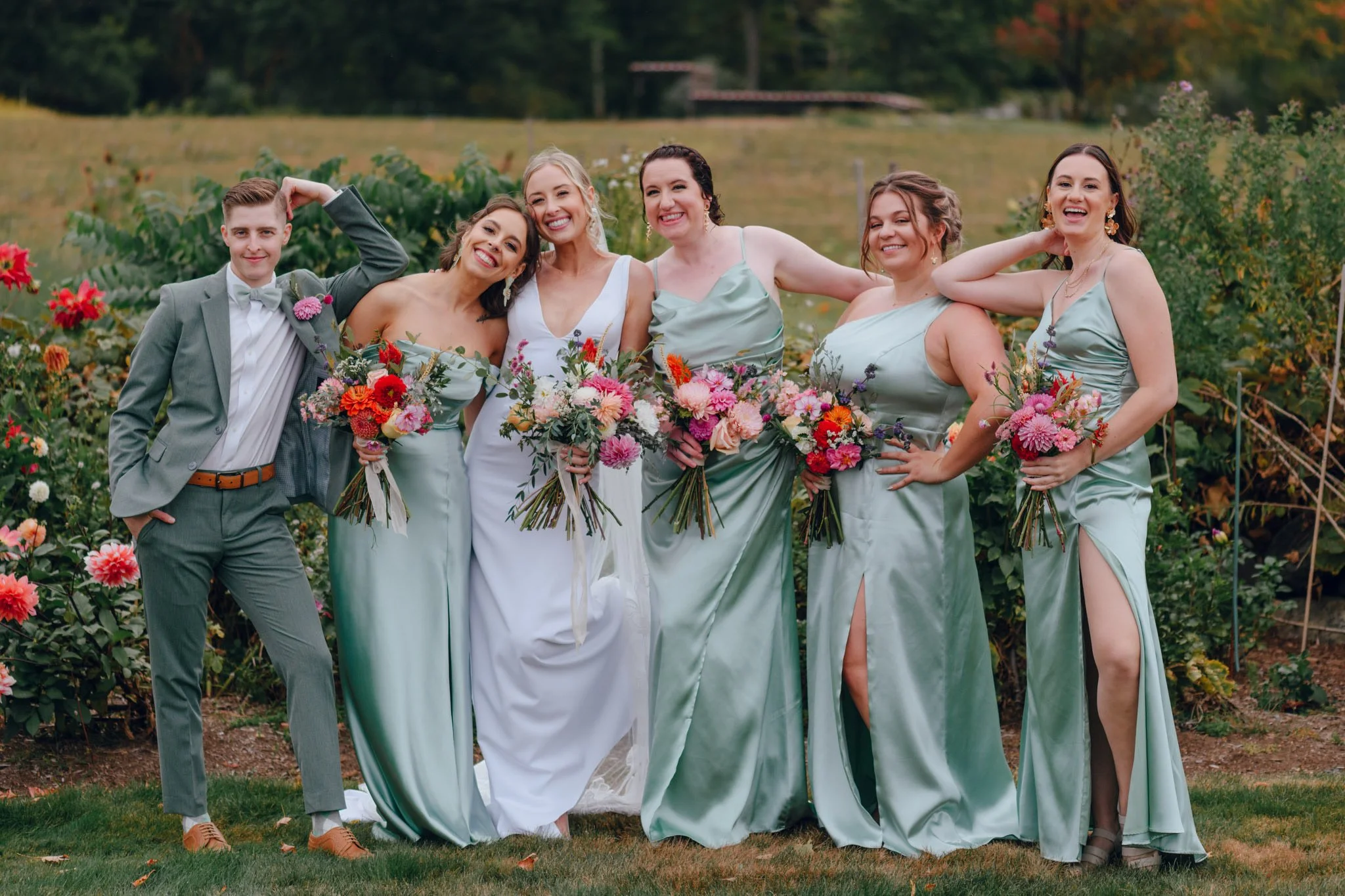 A group of six women and one man standing outdoors in a garden with flowers, dressed in wedding attire. The women are wearing sage green bridesmaid dresses, and the bride is wearing a white gown. The man is in a light gray suit. They are smiling and 