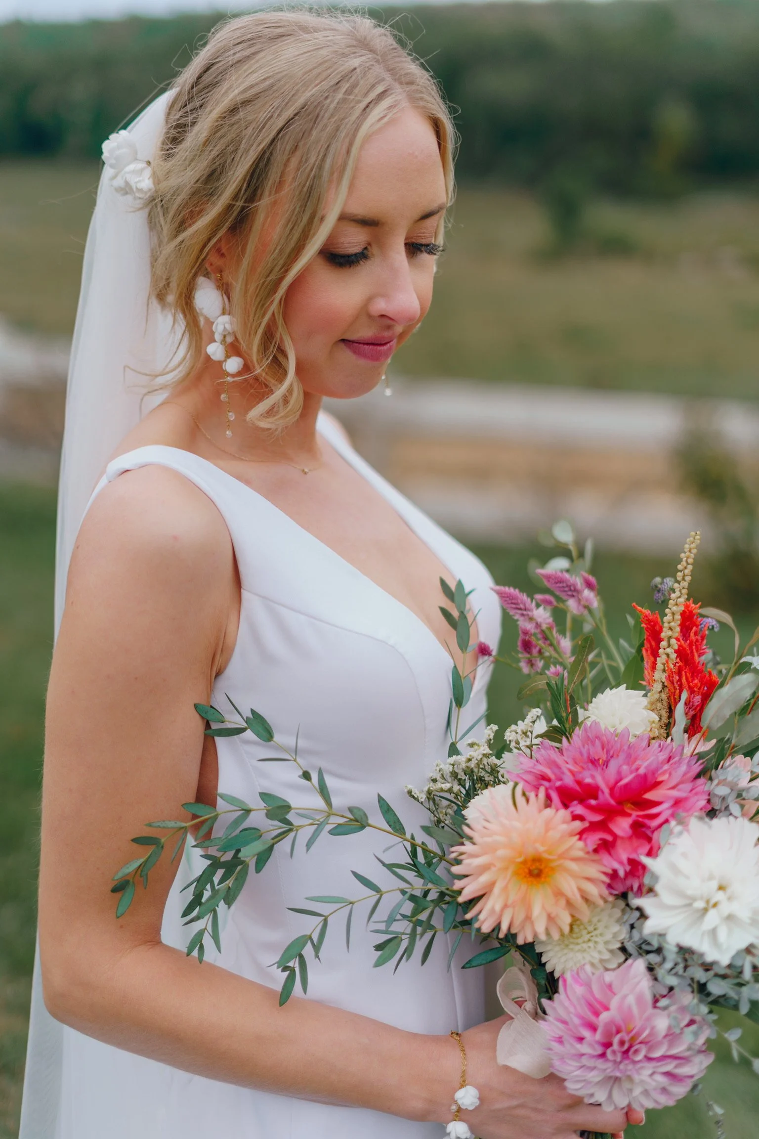 A bride in a white dress holding a bouquet of pink, white, and red flowers with green foliage, standing outdoors with a blurred landscape background.