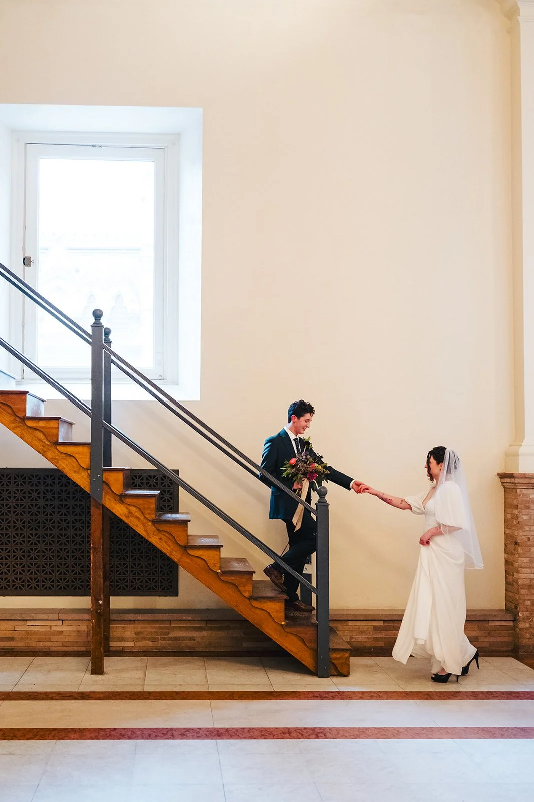 A bride in a white wedding dress and heels meets her groom on a staircase inside a building, holding hands and smiling.
