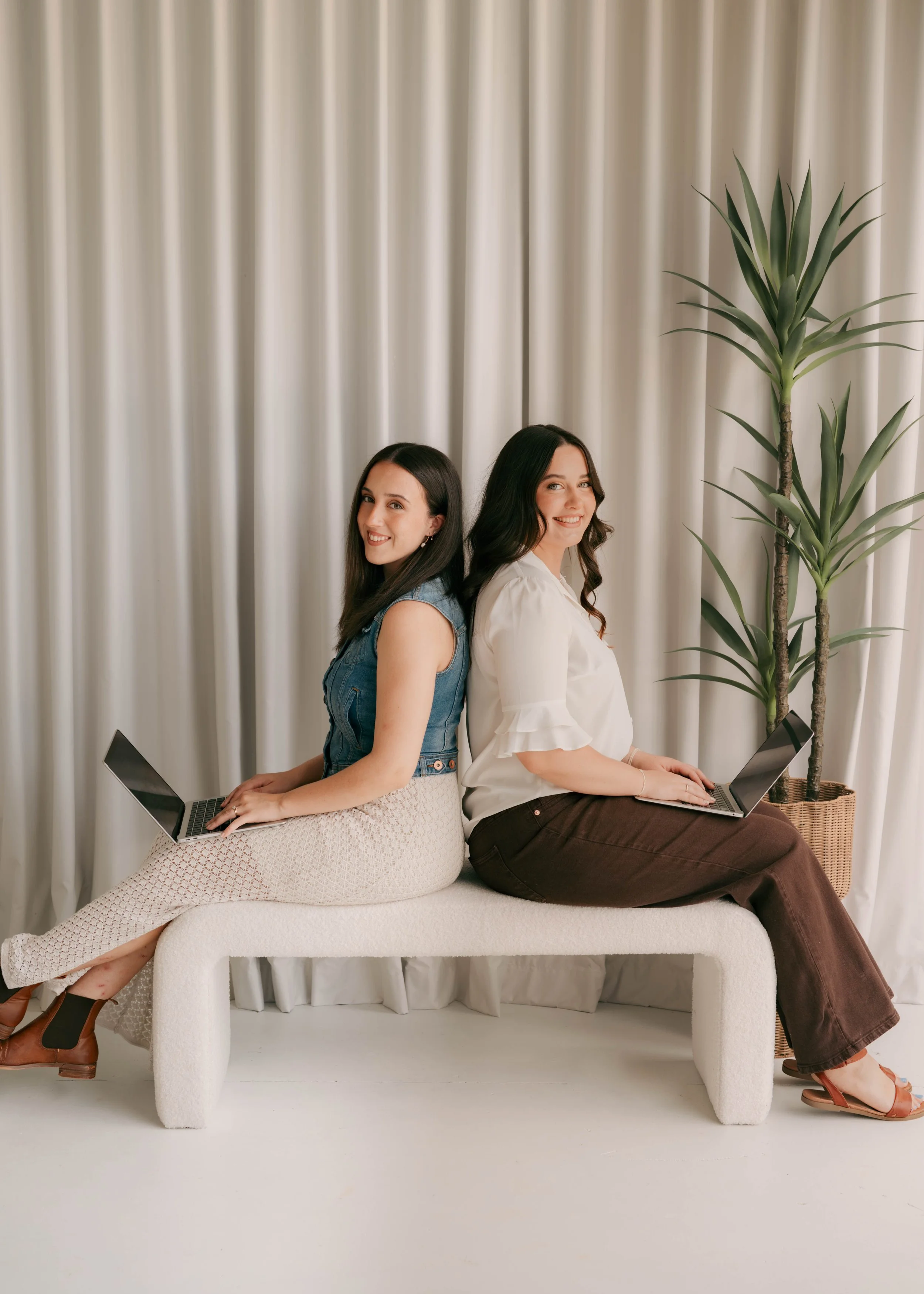 Two women sit back-to-back on a white modern sofa, each working on a laptop. They are smiling and looking at the camera. The woman on the left has dark hair and is wearing a sleeveless denim top with beige pants. The woman on the right has wavy dark 