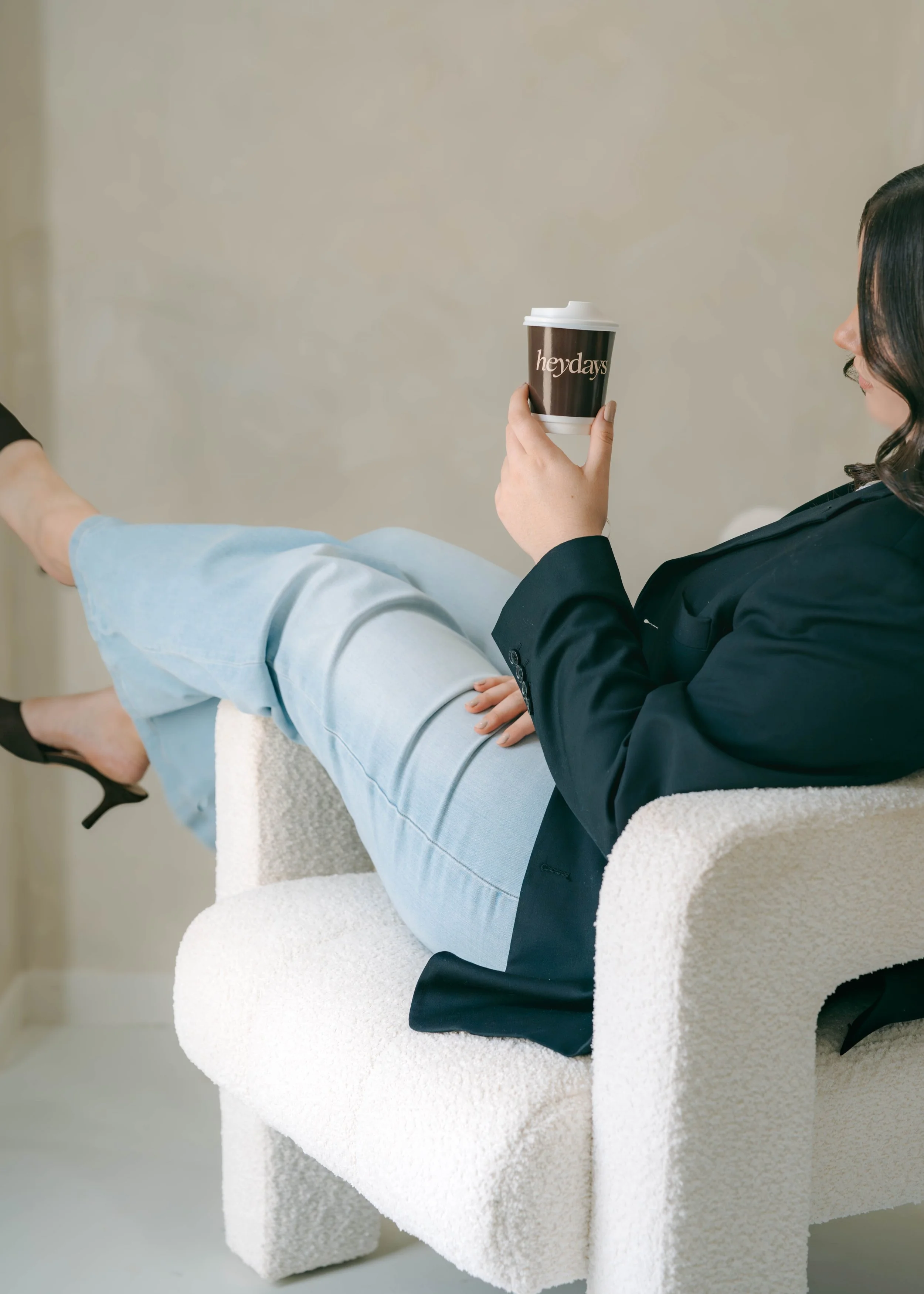 A person sitting on a plush white chair with their legs crossed, holding a Starbucks cup with "hey days" on it, wearing black blazer and light blue jeans, with black high heels, in a minimalistic room with beige walls.