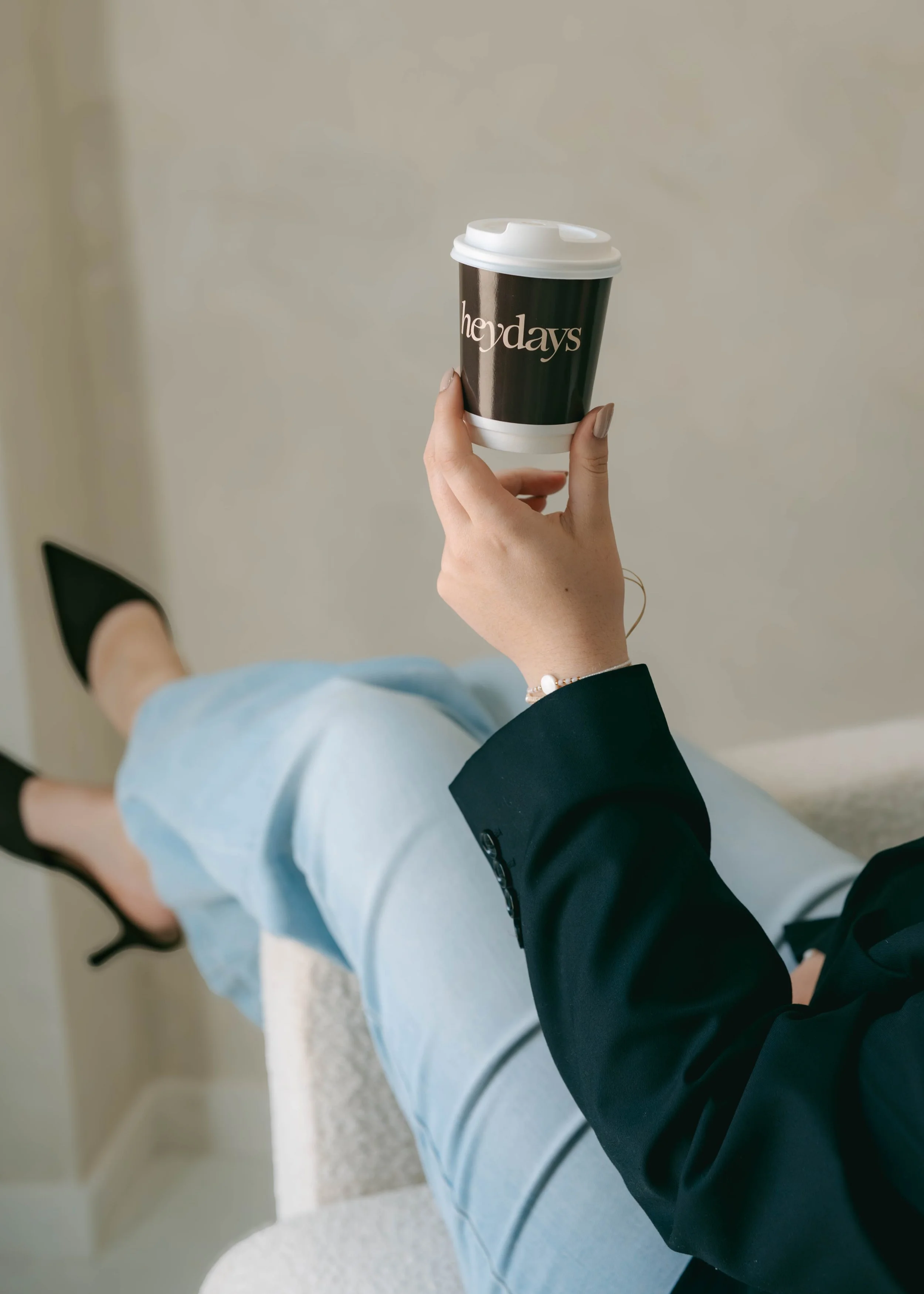 A person sitting in a chair with their legs crossed, holding a coffee cup labeled 'her days' in one hand. The person is dressed in a black blazer, light blue jeans, and black shoes, and is wearing a bracelet on the wrist. The background is neutral, a