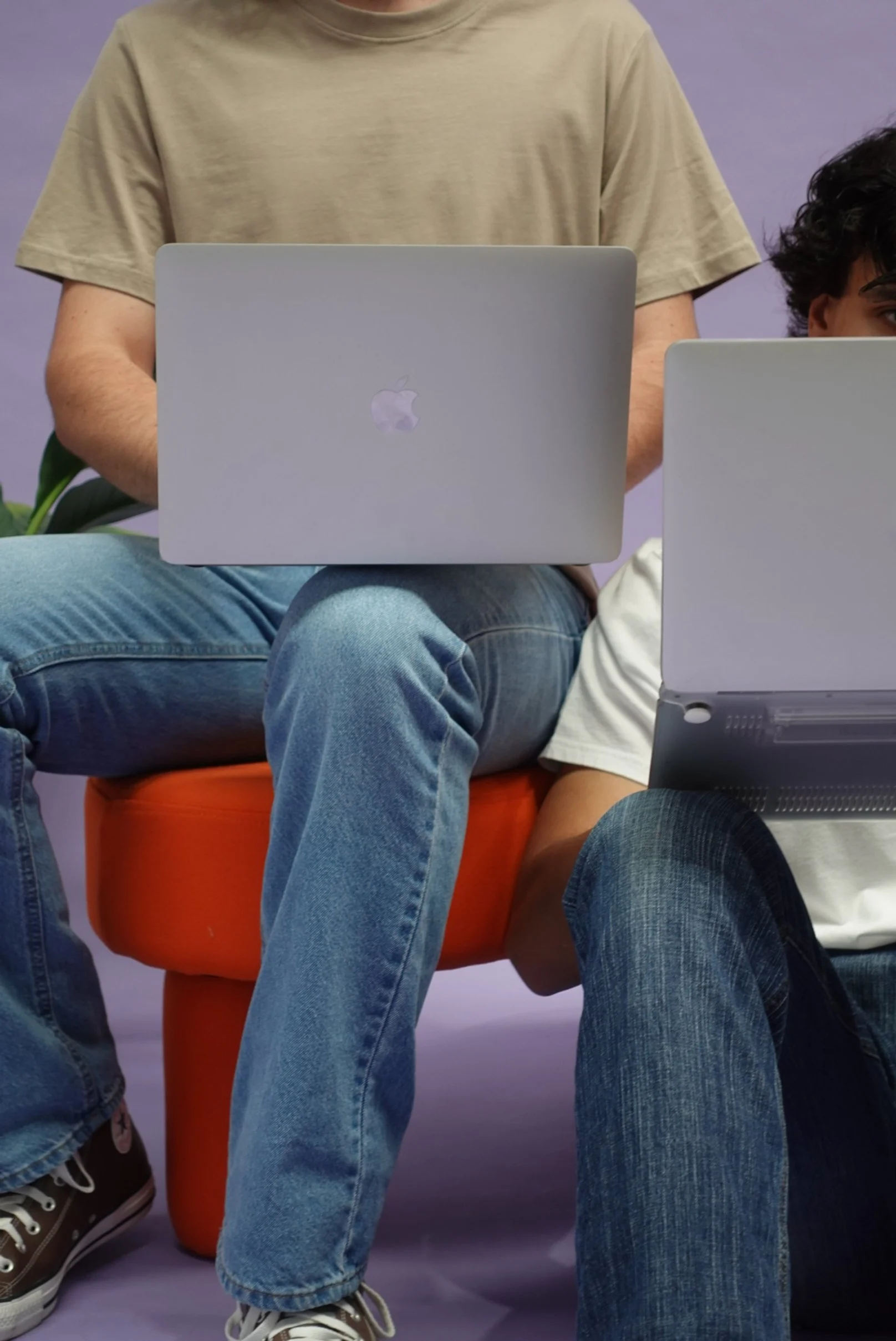 Two individuals sitting cross-legged on an orange stool, each using a silver Apple MacBook laptop, with a purple background.