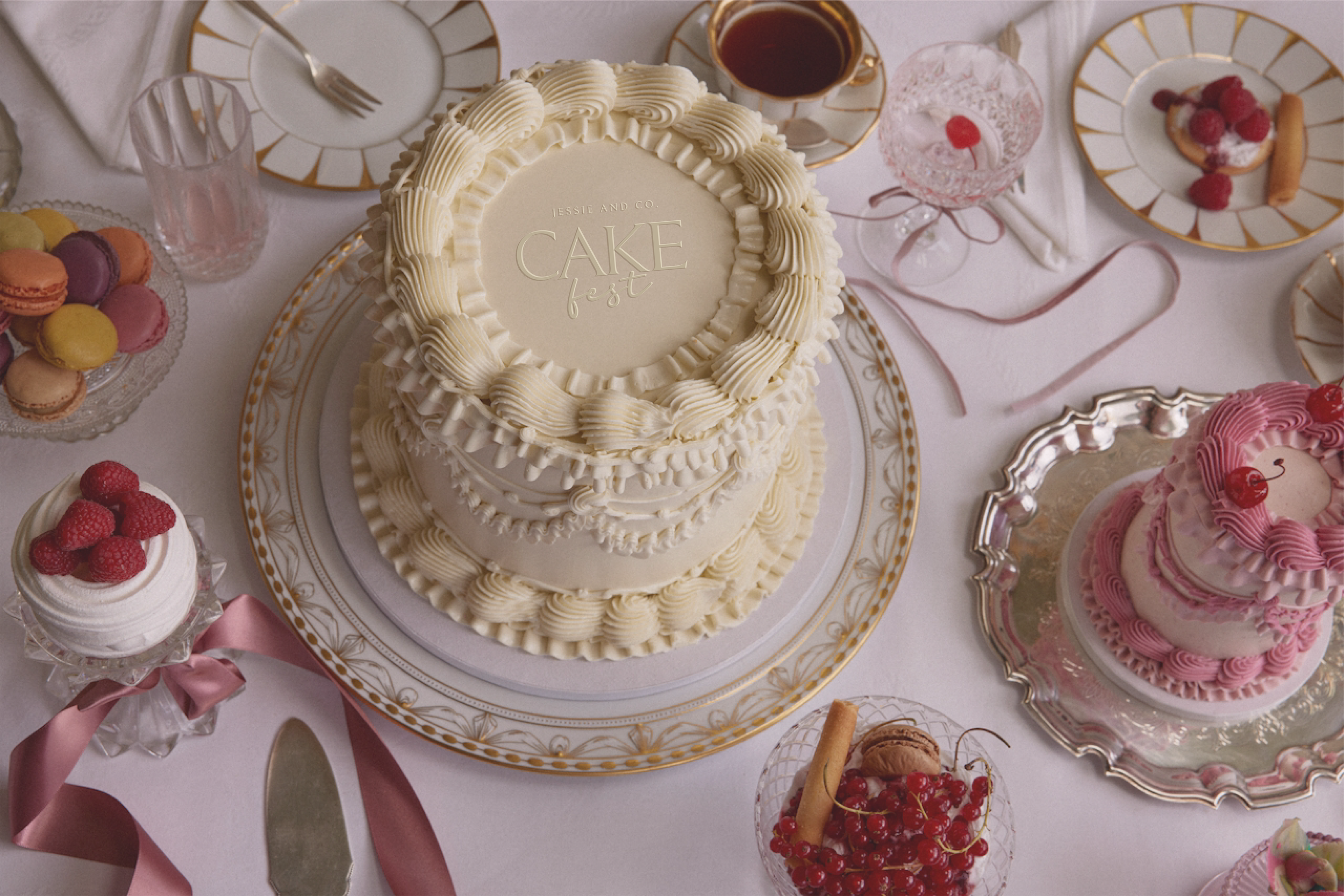 A decorated cake with the words "Cake Fest" written on it, topped with pastel-colored flowers, with a slice of the cake on a small plate beside it, decorated with flowers and greenery, against a light purple background.
