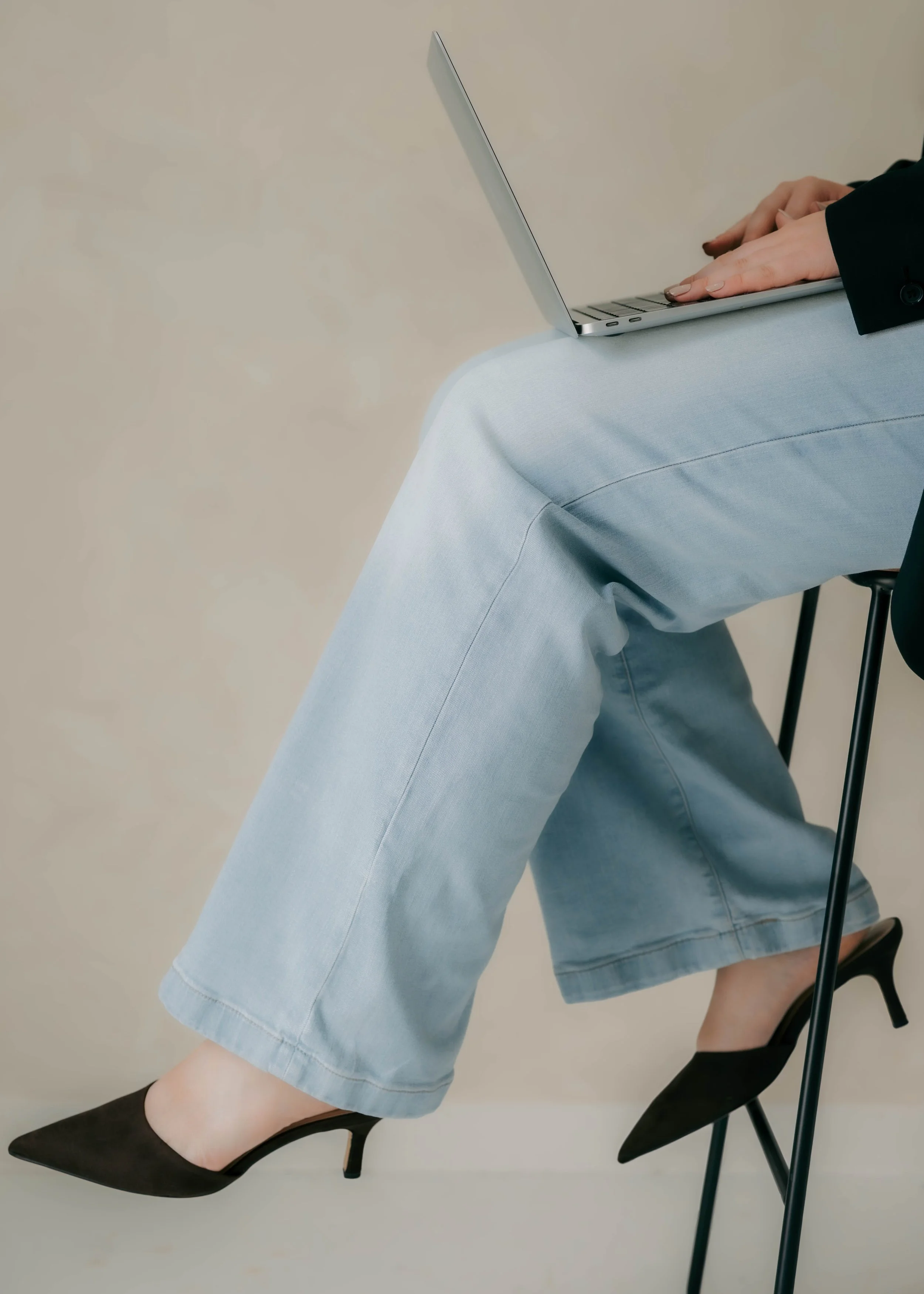 A person sitting on a black chair wearing black high heels, light blue jeans, a black blazer, with their hands on a silver laptop on their lap.