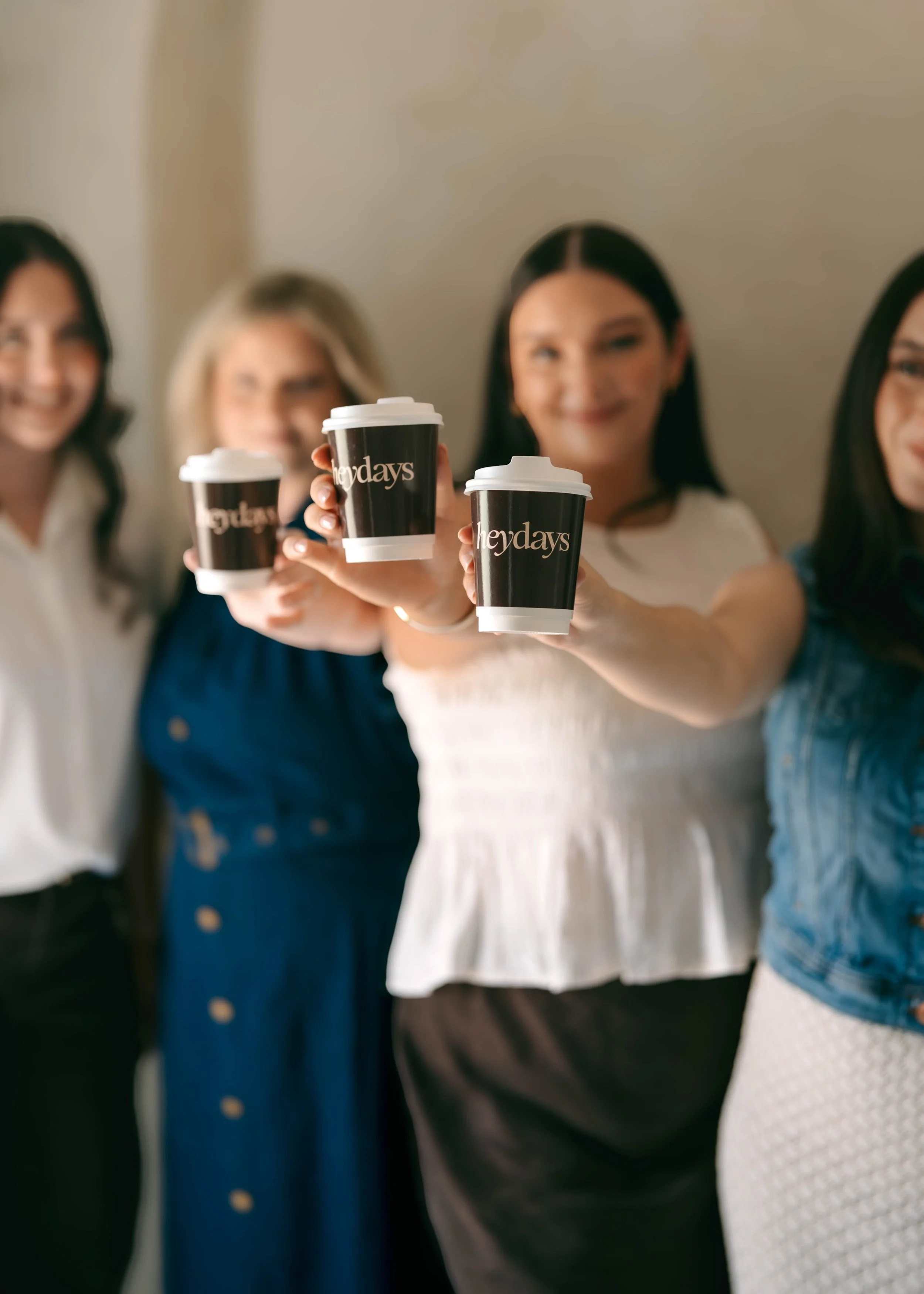 Four women holding coffee cups with 'hey days' logo, smiling at the camera.