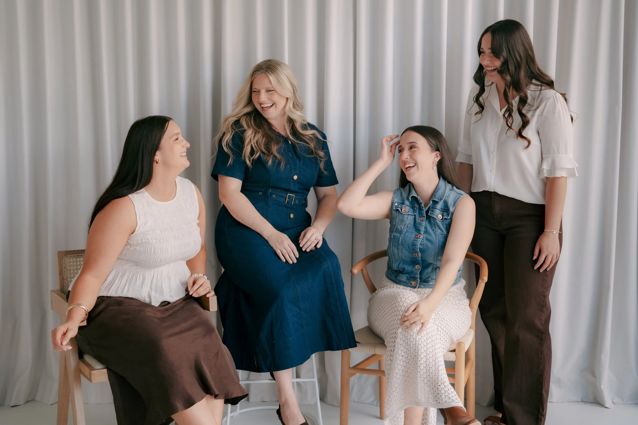 Group of five women laughing and talking in front of a white curtain, some sitting and some standing.