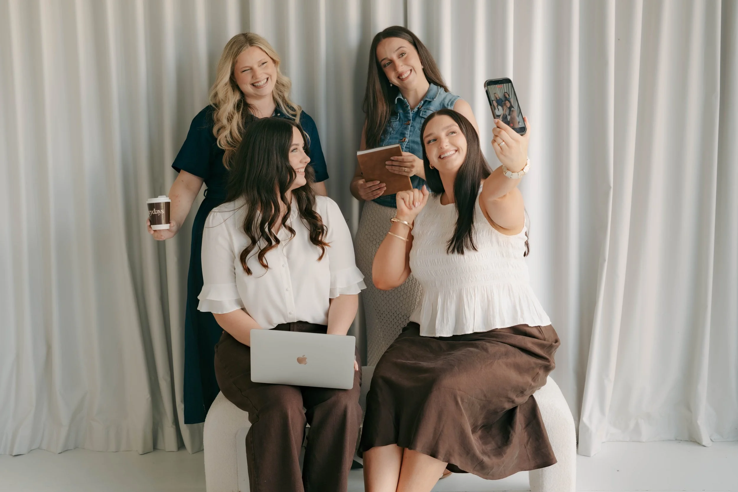 Four women taking a selfie together in a room with white curtains, seated and standing around a laptop, smiling and enjoying each other's company.
