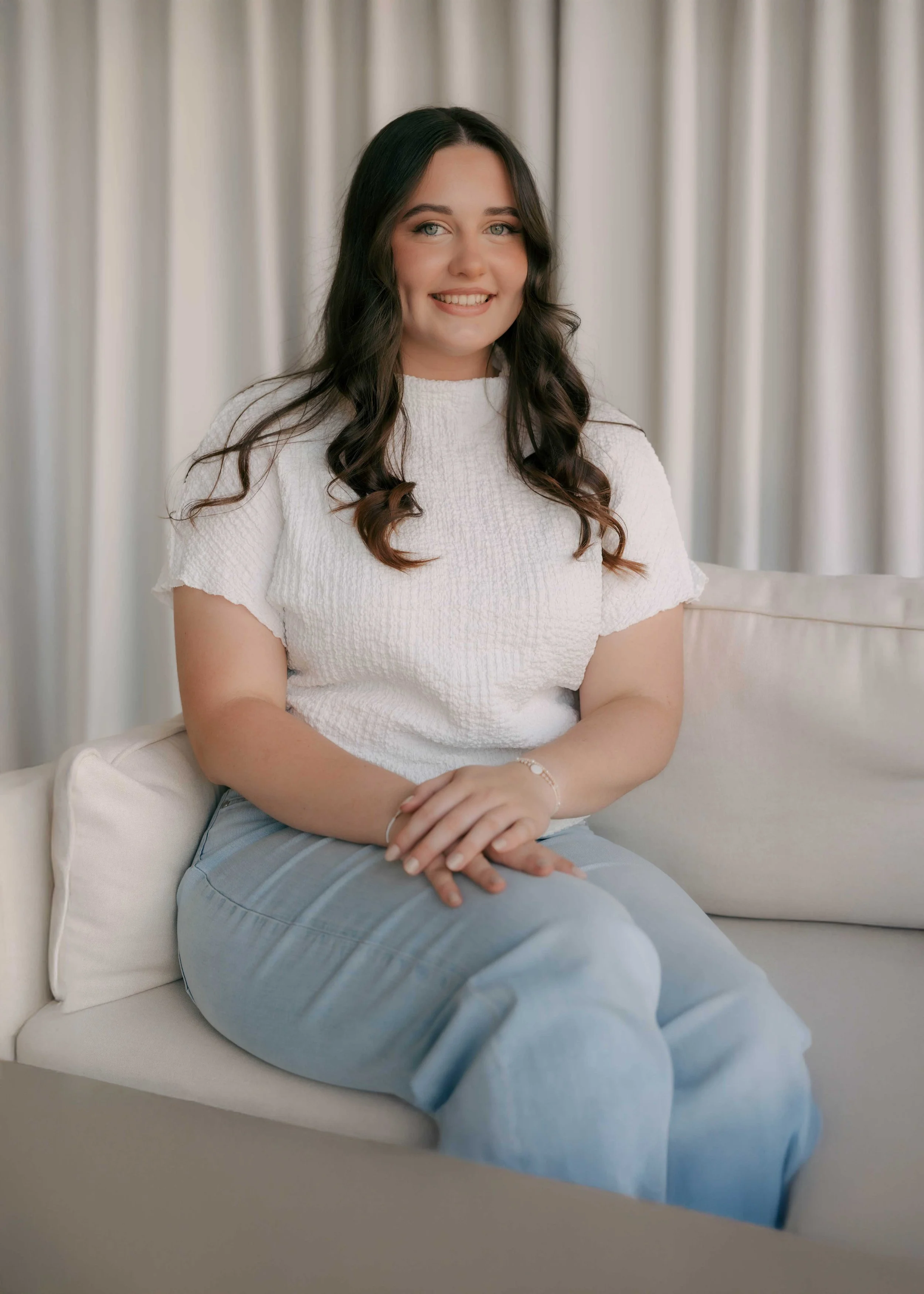 A young woman with dark wavy hair and blue eyes, smiling, sitting on a cream-colored sofa with white pillows, in front of beige curtains.