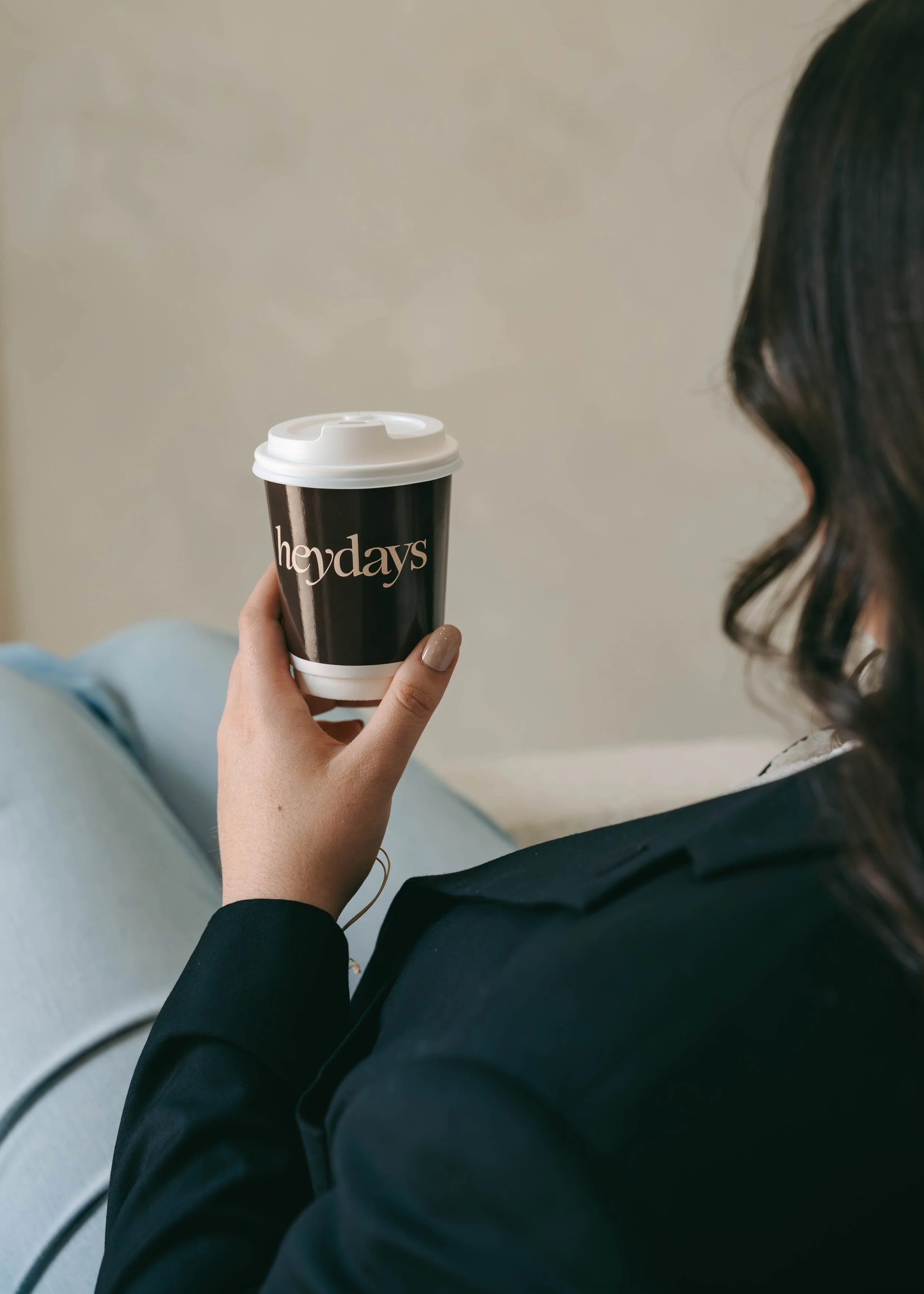 Person holding a black and white coffee cup with 'heyday' printed on it, sitting down, wearing a black jacket and light blue jeans, with dark hair visible on the right side of the image.