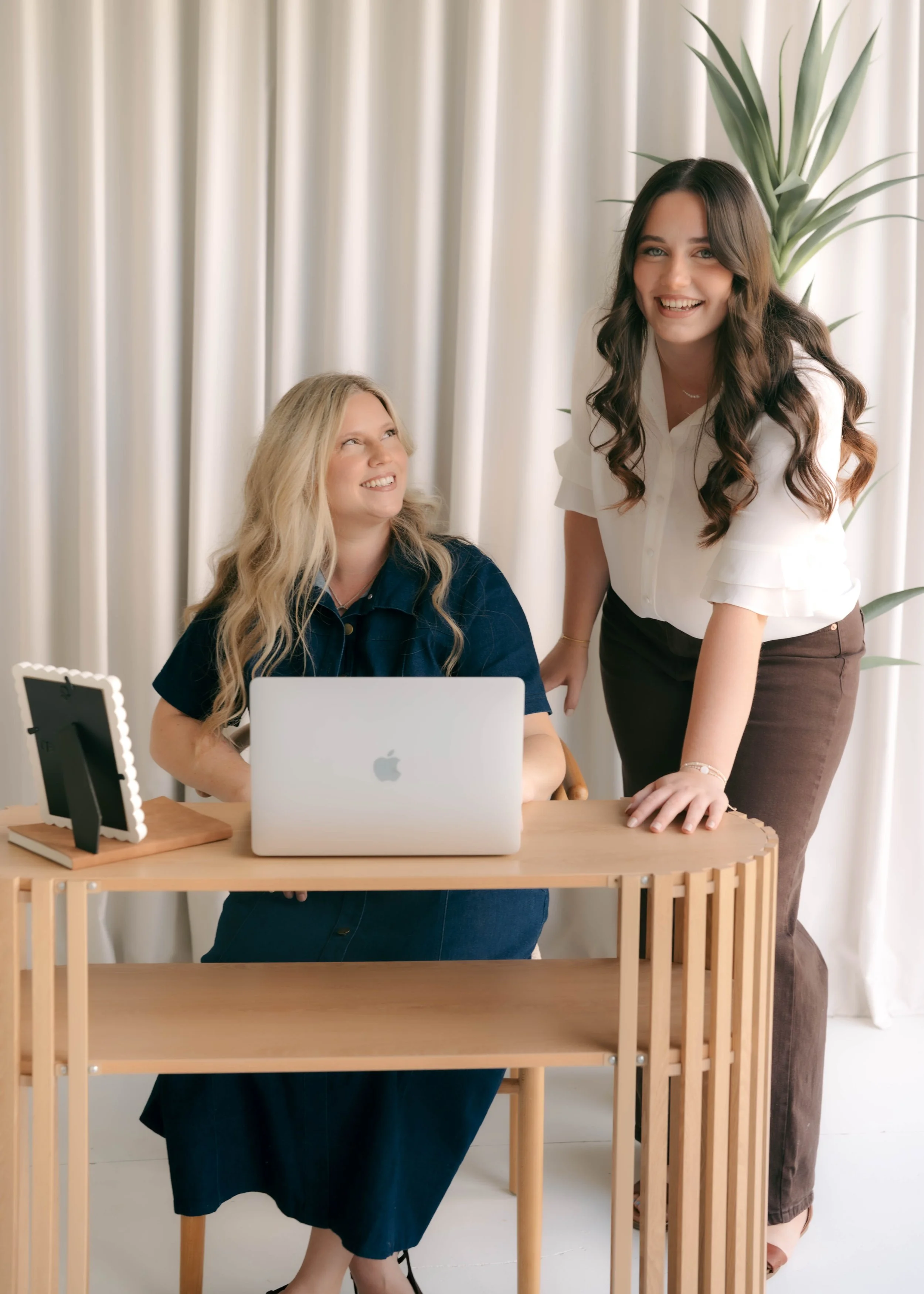 Two women at a desk with a laptop, one sitting and one standing, smiling and looking at each other in a bright room with white curtains and a large houseplant in the background.