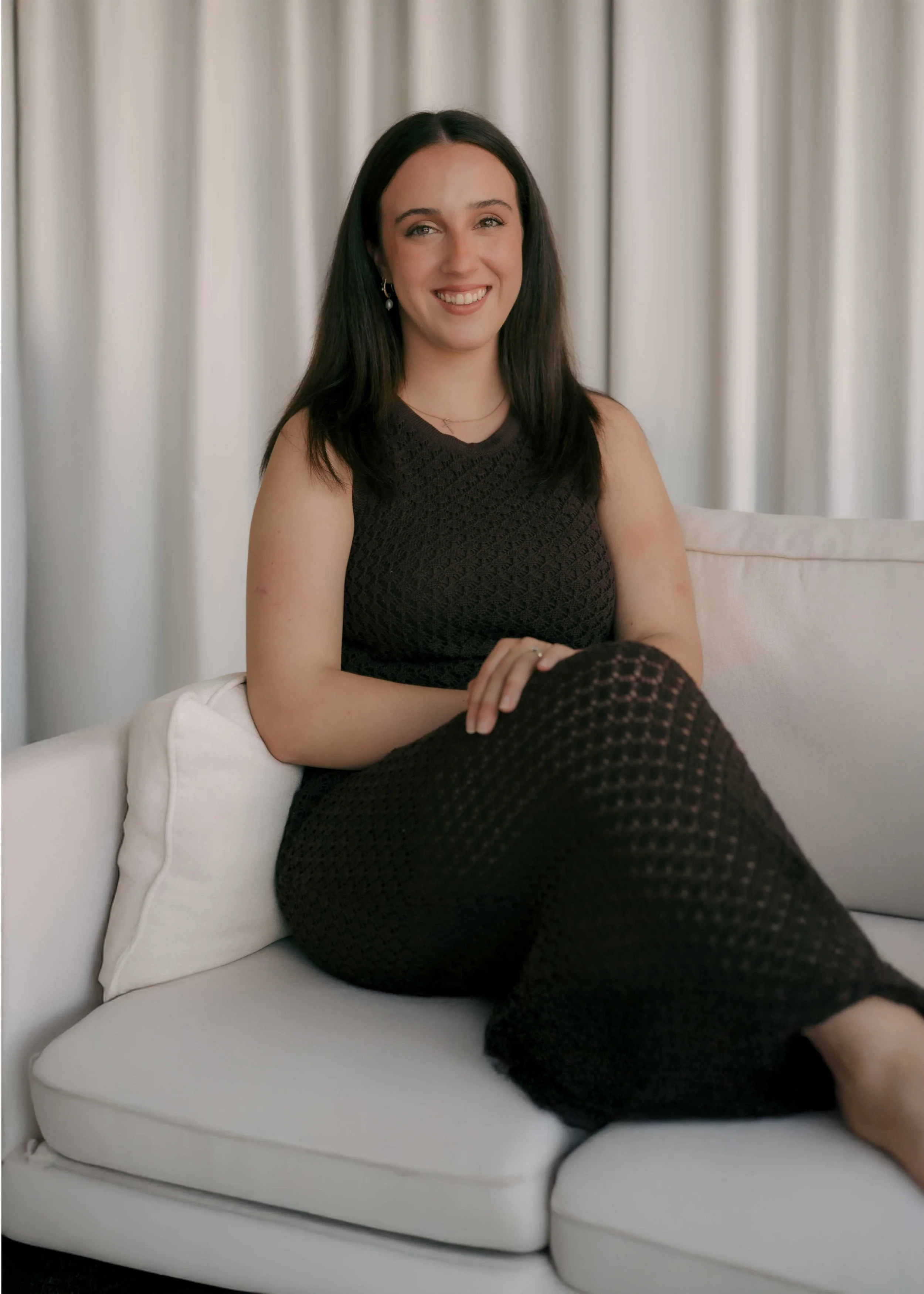 A woman with long dark hair sitting on a white couch, smiling with her hand on her knee, in front of white curtains.