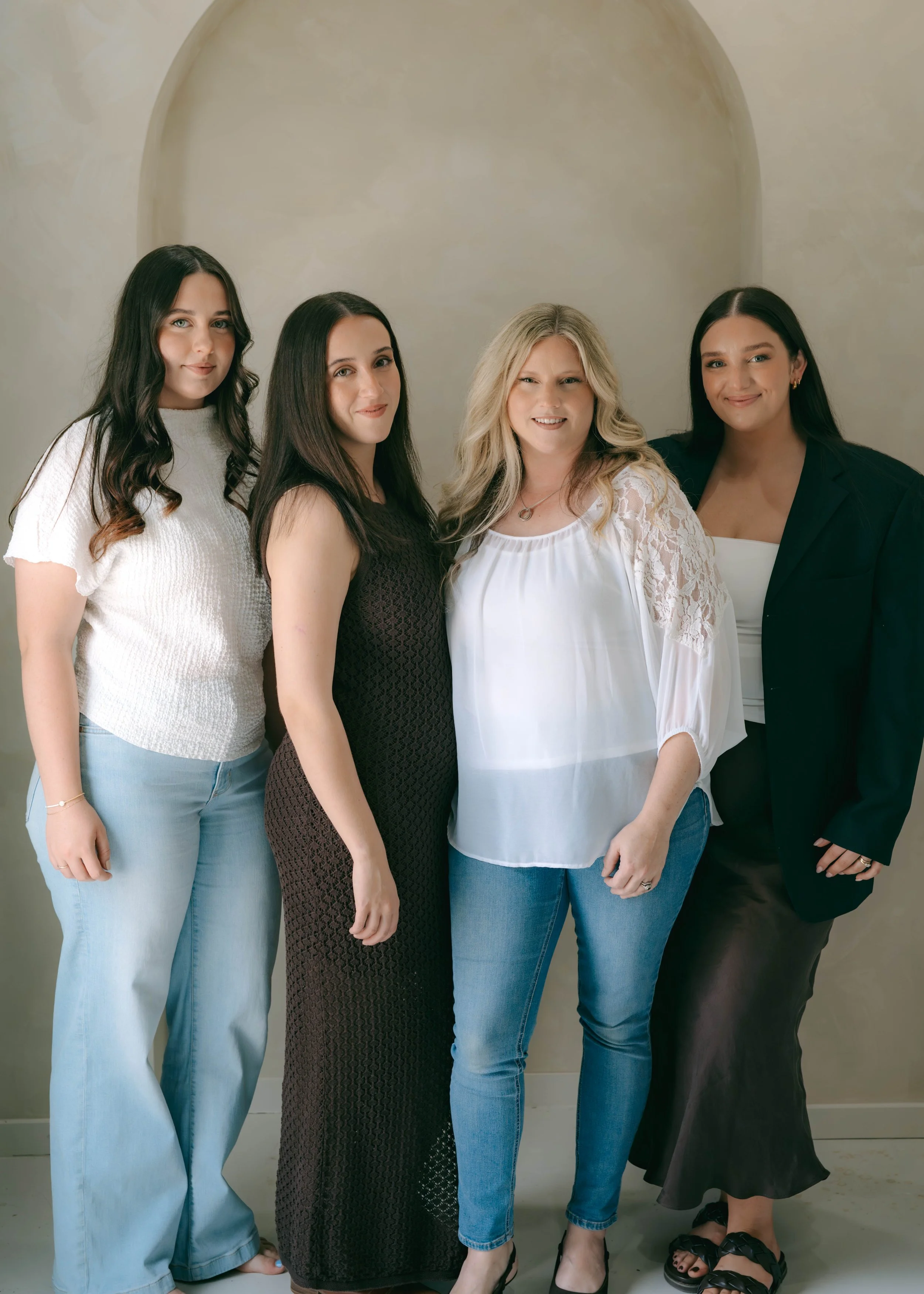 Group of four women standing close together against a neutral background, smiling, dressed in casual and semi-formal attire.