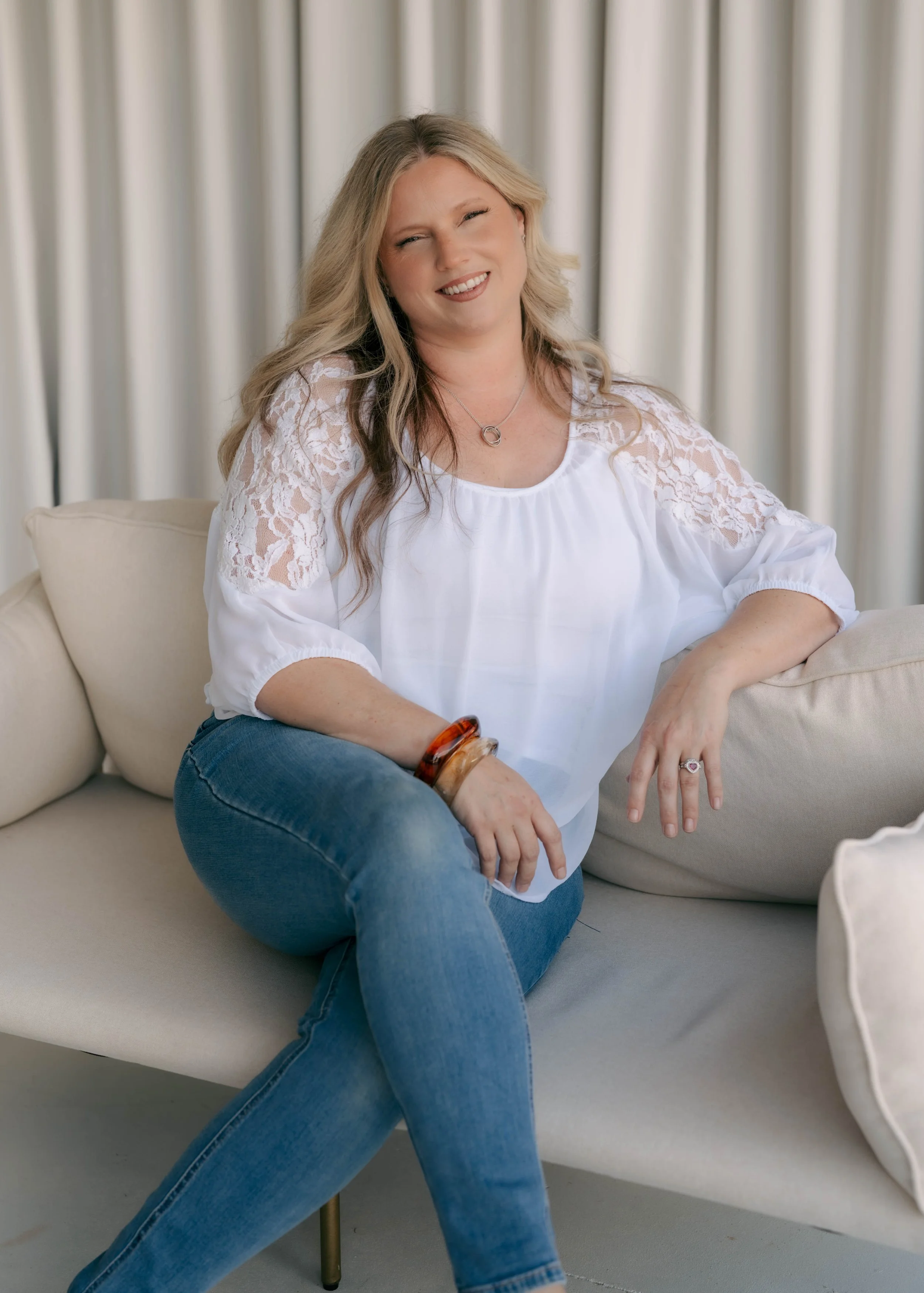 A woman with long blonde hair sitting on a beige sofa, smiling, in front of cream-colored curtains, wearing a white blouse with lace sleeves and blue jeans.