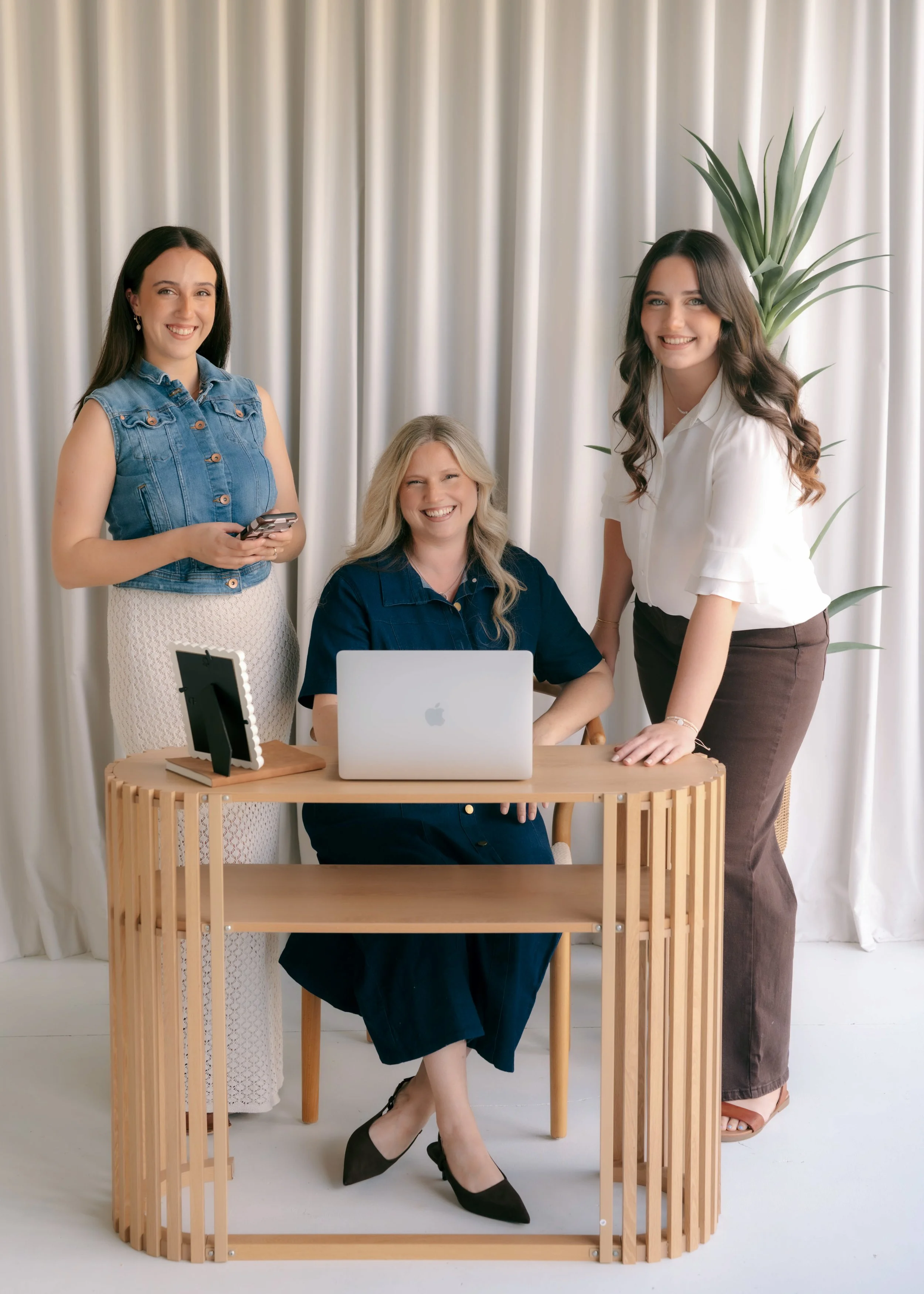 Three women in an office setting with a white curtain background, one sitting at a wooden desk with a silver laptop, the other two standing beside her, smiling and posing for the photo.
