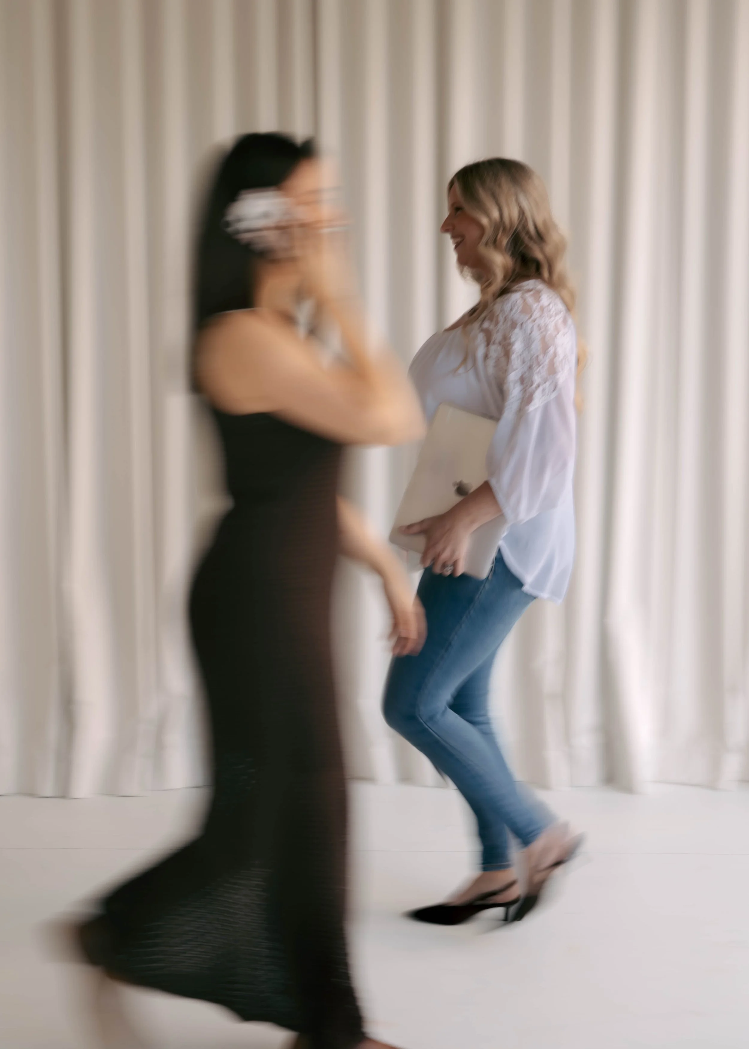 Two women dancing in front of a cream-colored curtain, with motion blur indicating movement. One woman is wearing a black dress, glasses, and a necklace, while the other is wearing a white lace top and blue jeans, holding a notebook or tablet.