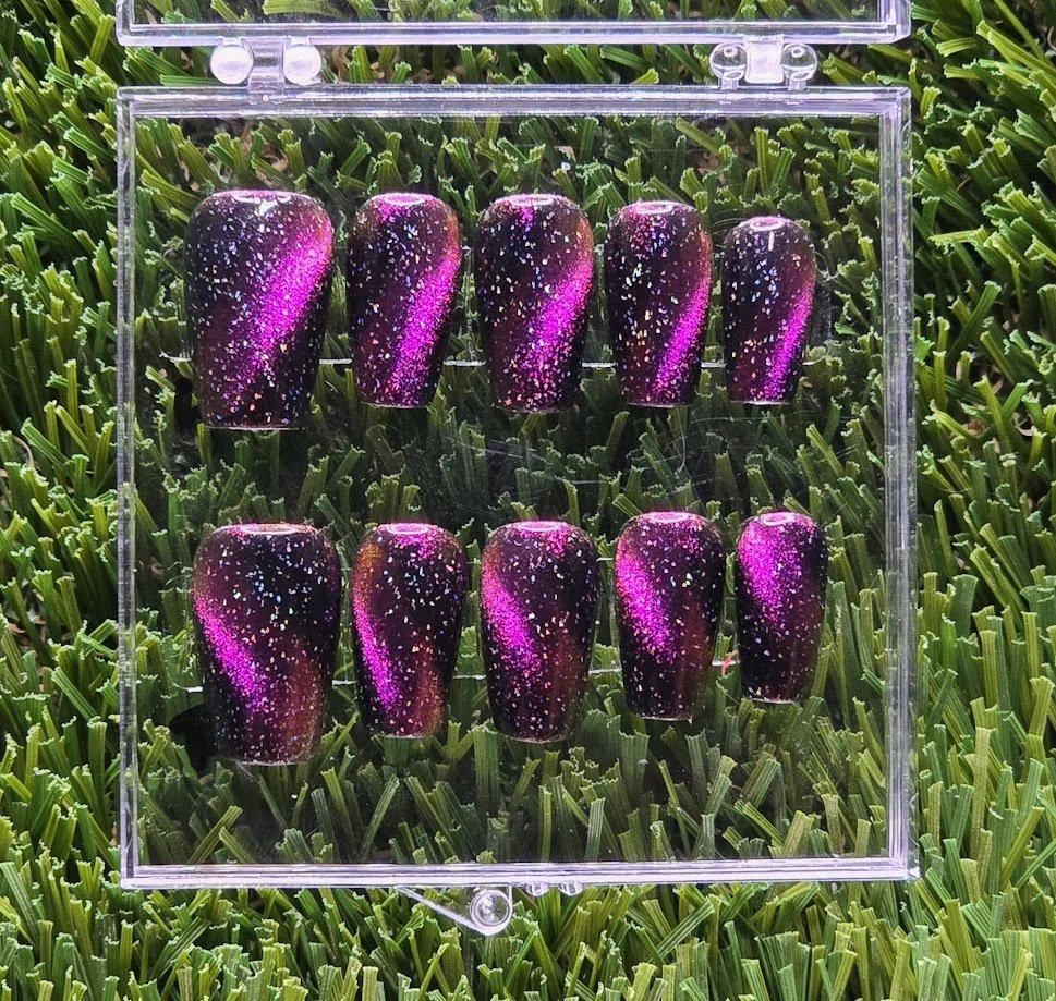 Display case with ten galaxy-themed artificial nails painted with purple, black, and white speckled designs, on green grass background.