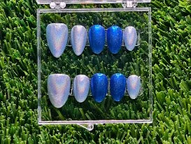 A set of artificial nails painted in various shades of blue, displayed on a clear acrylic stand outdoors on grass.