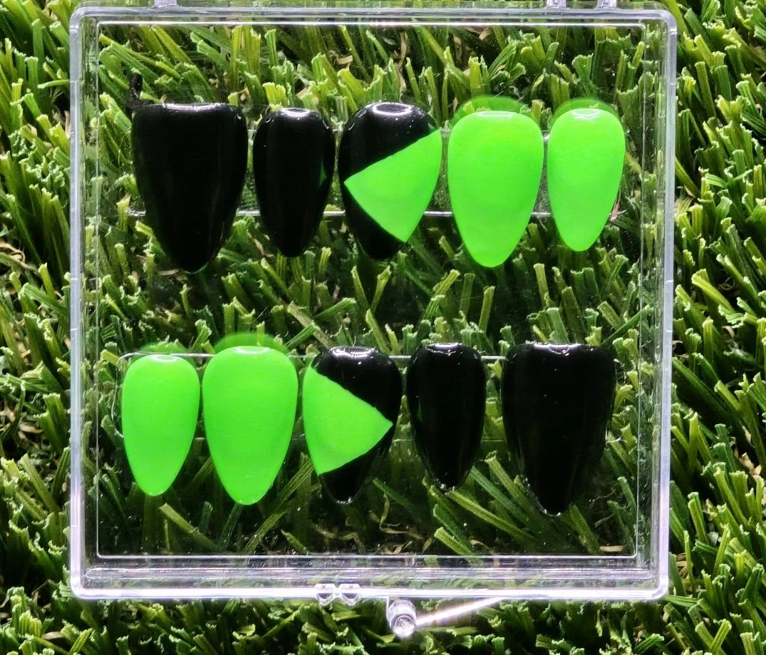 A set of ten artificial nails in black and bright green, arranged in two rows in a clear plastic case, placed on a bed of green grass.