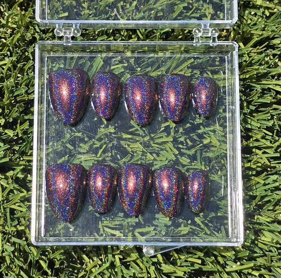 A transparent plastic box holding ten glittery, holographic, dark-colored stones arranged in two rows on green grass.