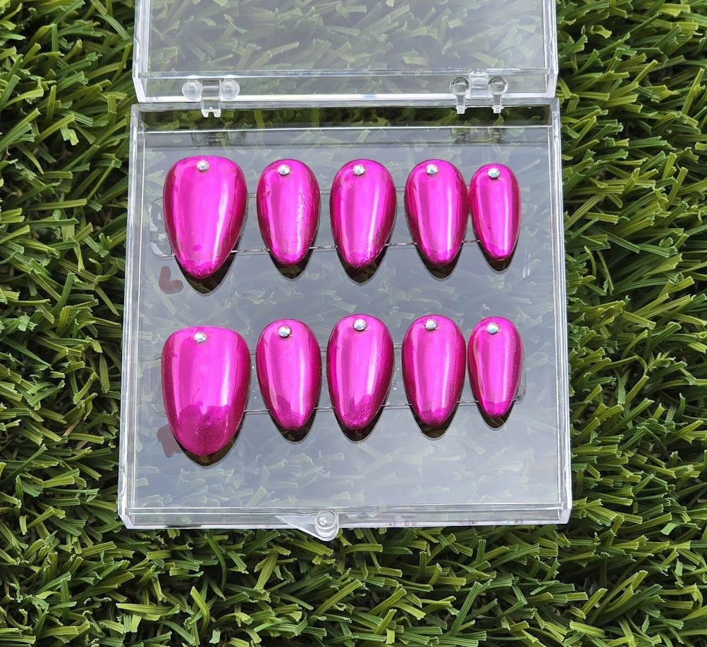 A plastic box containing ten shiny pink teardrop-shaped accessories with small rhinestones on top, placed on green artificial grass.