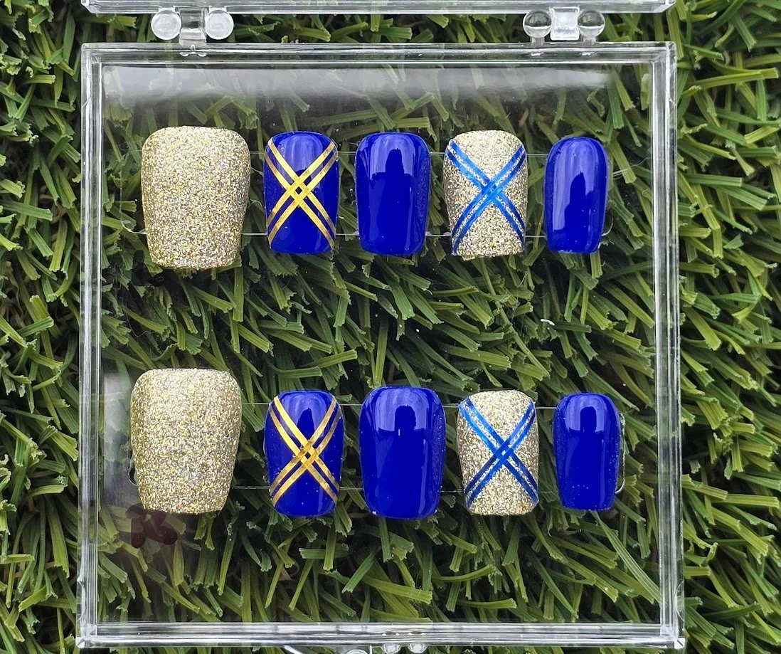Set of ten artificial press-on nails arranged in two rows inside a clear acrylic box, with alternating designs of gold glitter and royal blue with gold and blue diagonal line patterns, on a grassy background.