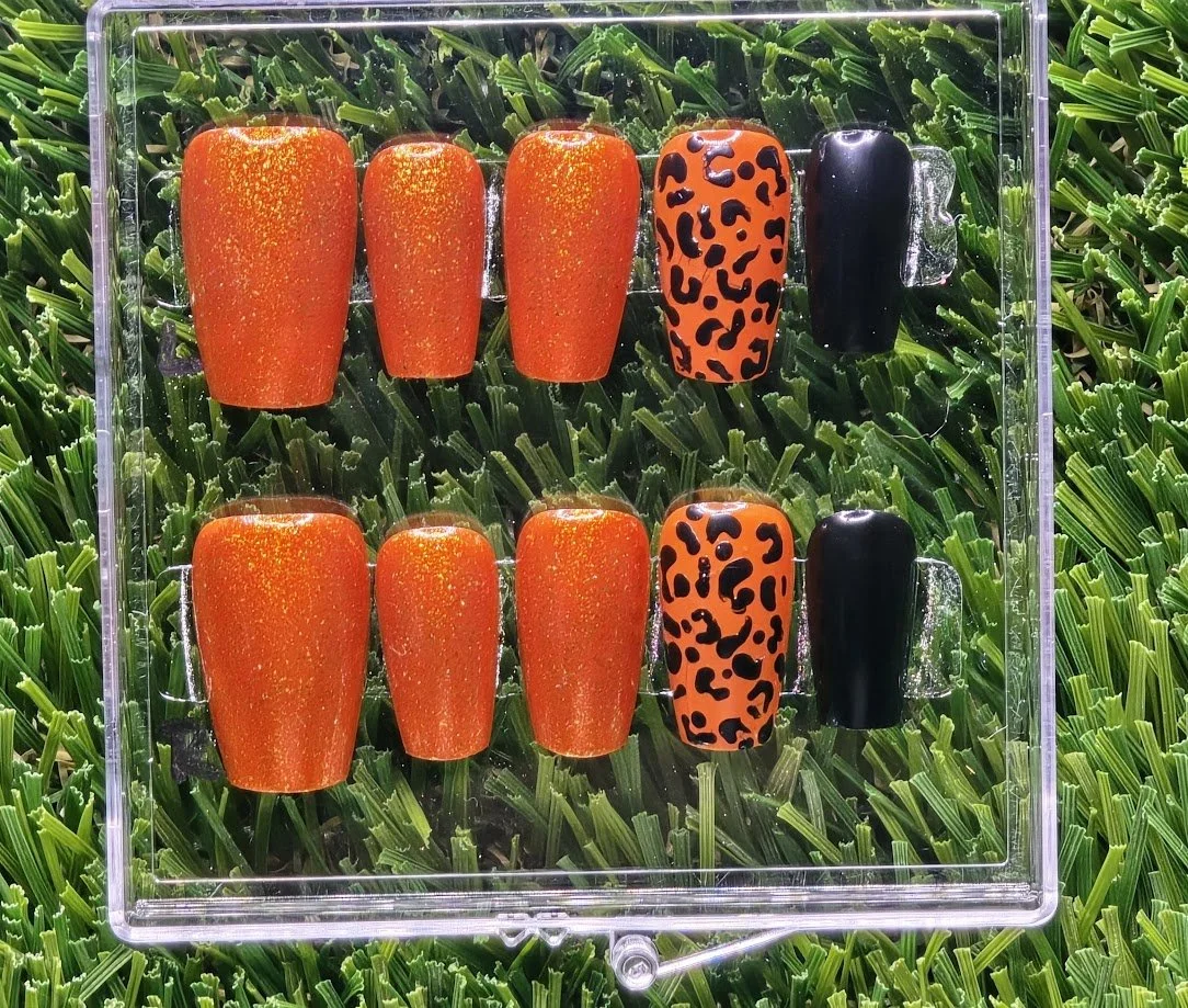 Set of ten orange and black animal print and solid black false nails arranged in a display case on green foliage background