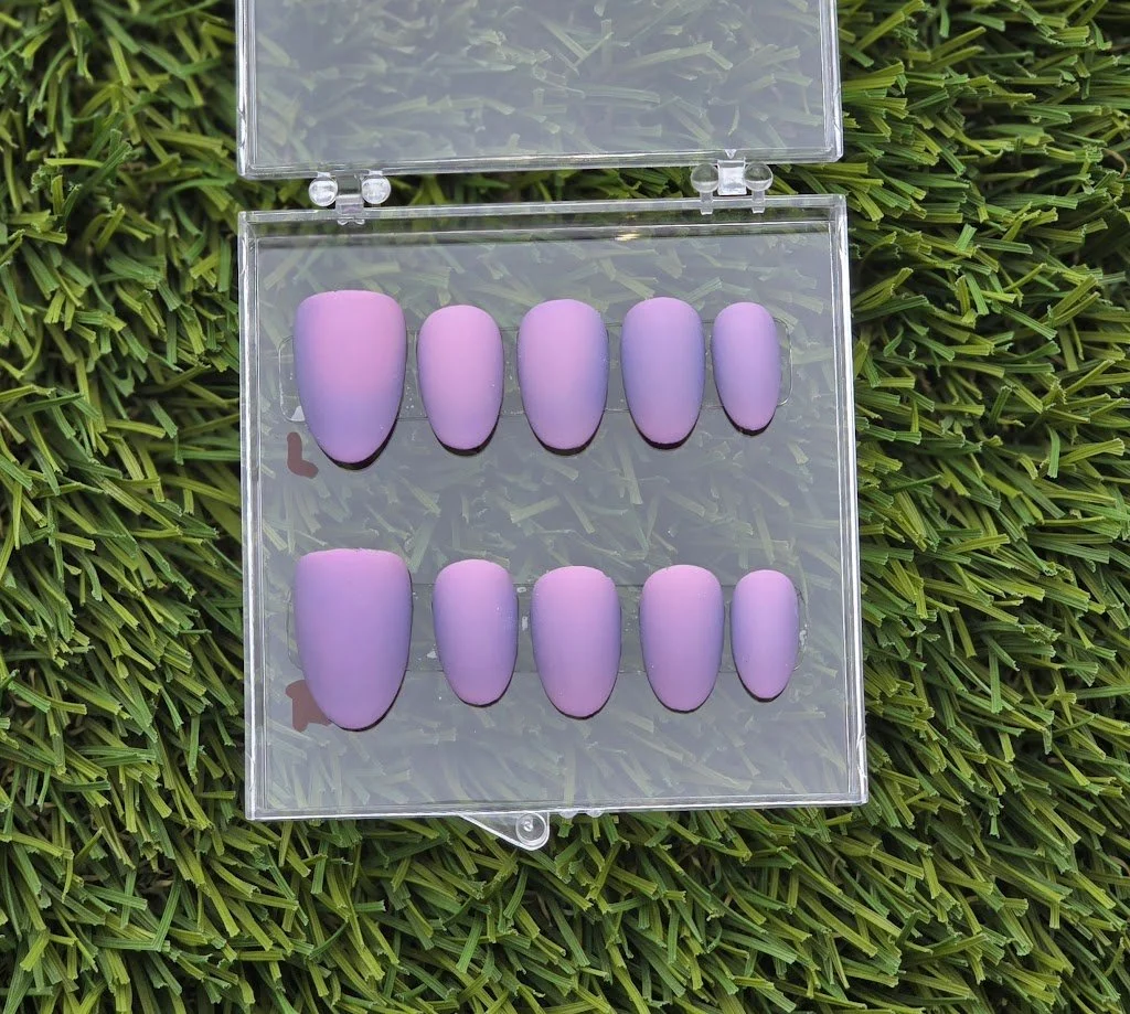 Set of artificial purple nails in a clear plastic case on green grass.