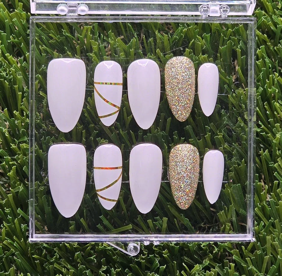 Set of ten nail art display tips with various designs, including plain white, white with holographic stripes, and glitter accents, arranged in a clear plastic case on a grass background.