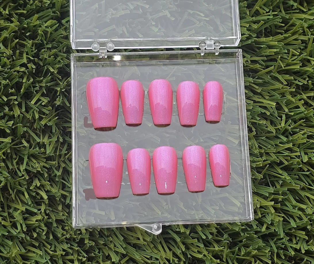 A set of ten pink artificial nails stored in a transparent plastic case on grass.