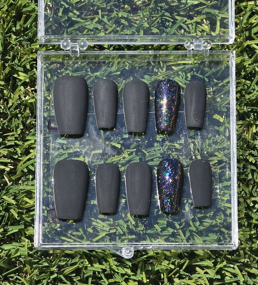 A clear plastic case with black press-on nails, some with multi-colored glitter, arranged on green grass.