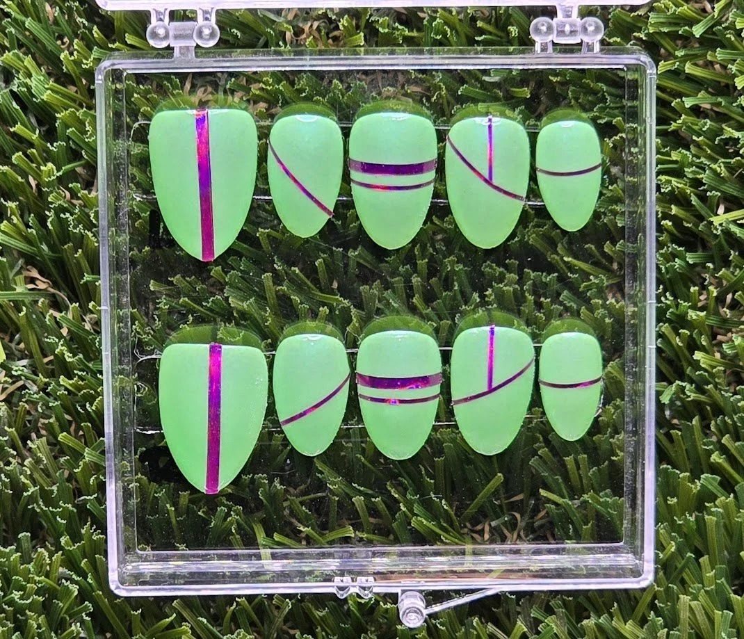 Collection of artificial green nails with purple designs, displayed in a clear plastic case on grass.
