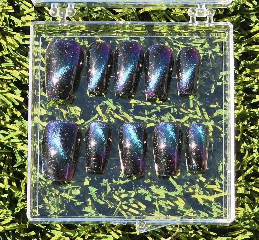 Nail tips painted with galaxy-themed artwork, featuring a dark background with blue, purple, and pink sparkles and stars, arranged in a clear plastic case on artificial green grass.