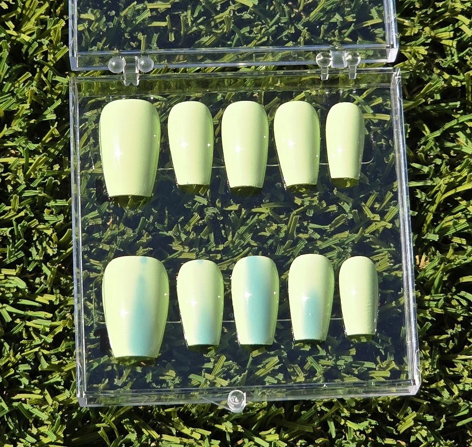 A clear plastic case with ten light green artificial nails inside, arranged on a grass surface.