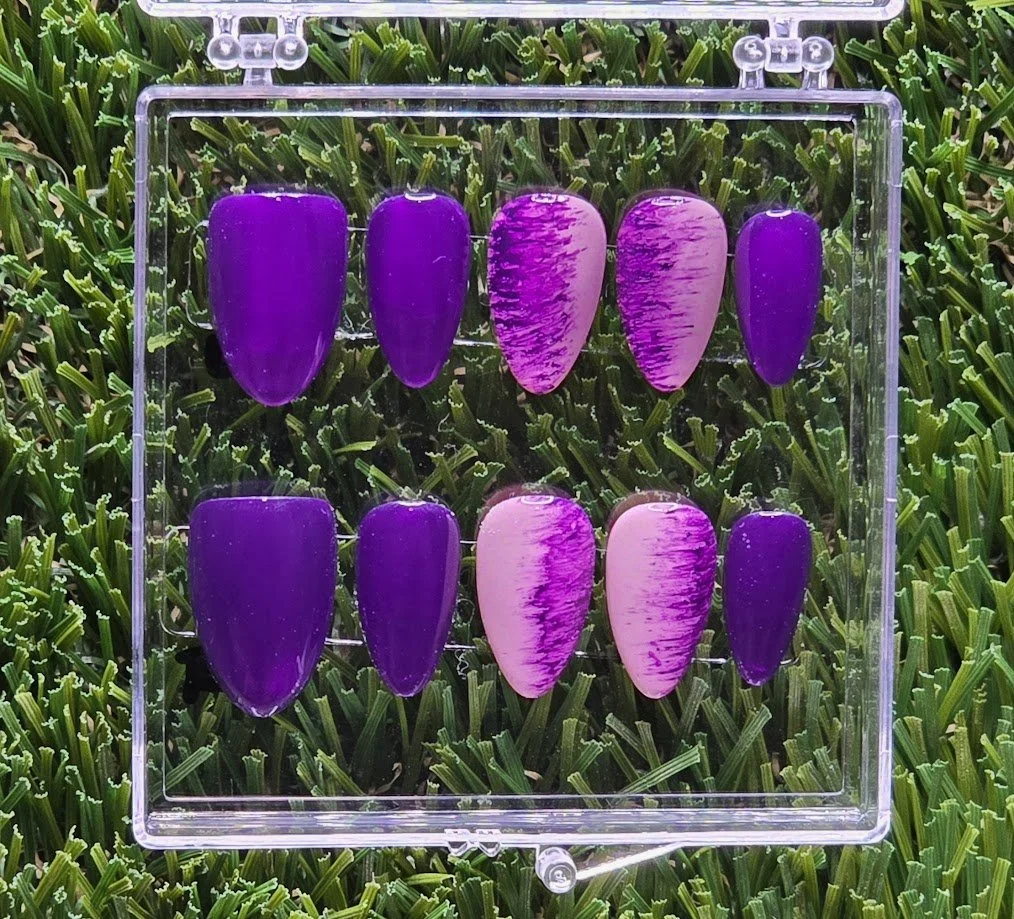 A display case containing ten purple and pink acrylic false nails arranged in two rows on a clear plastic stand, set on a bed of green grass.