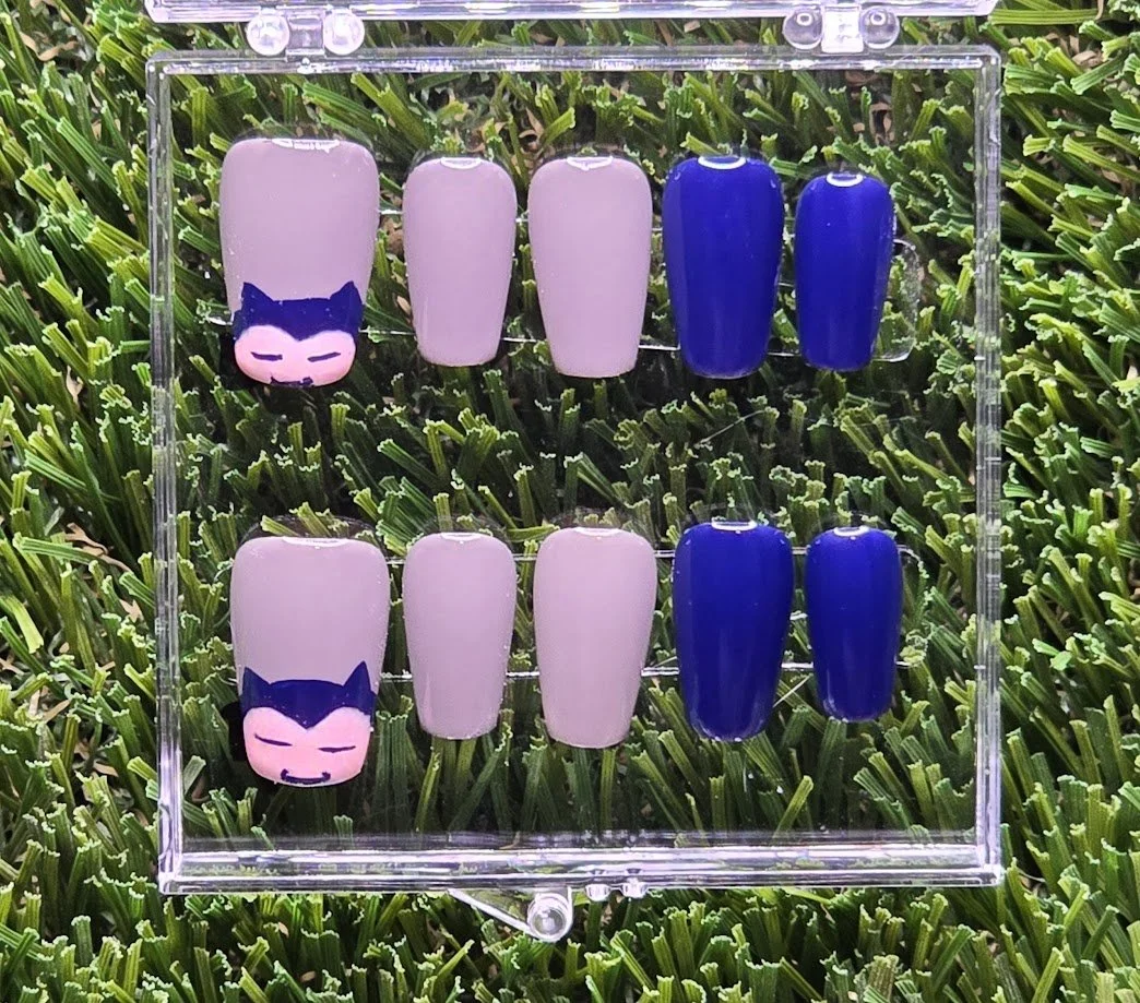 Set of ten artificial nails with purple, pink, and blue designs, displayed on a clear acrylic stand against a grassy background.