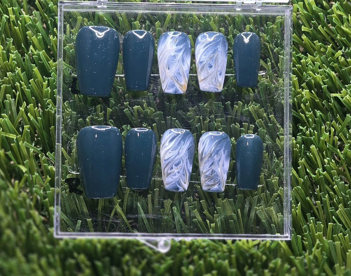 A set of ten artificial nail tips with blue and white marbled and solid dark blue designs, displayed in a clear plastic case on a grass background.