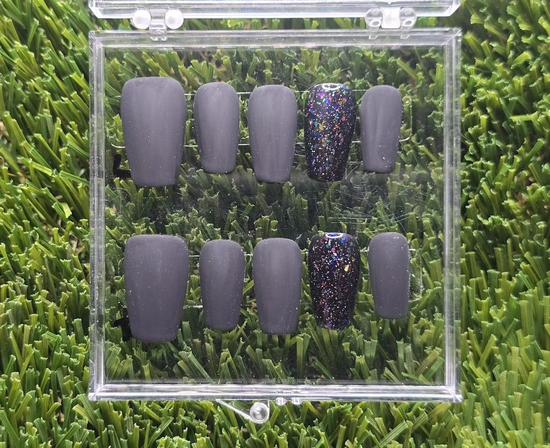 A set of matte gray and dark gray nail tips with two glittery black designs inside a clear plastic case, placed on green artificial grass.