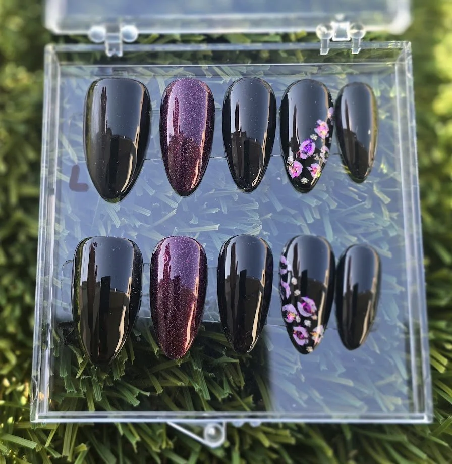 Set of ten black and purple gel nail polish swatches with floral and glitter designs in a transparent case on grass.